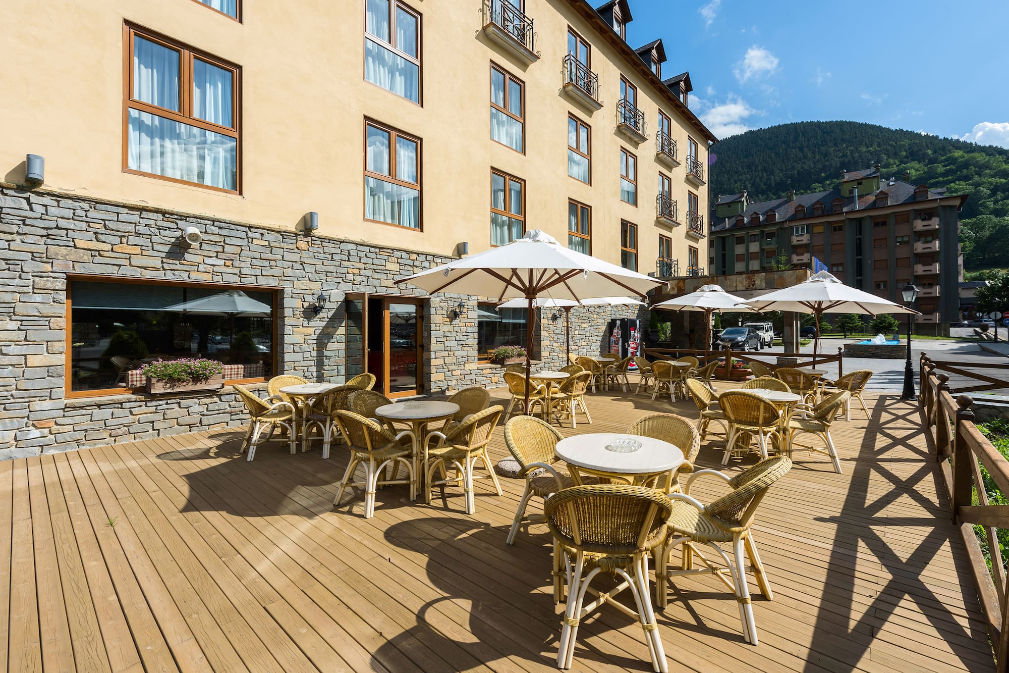 Terraza de hotel con mesas y sillas, parasoles, fachada de piedra y montaña.