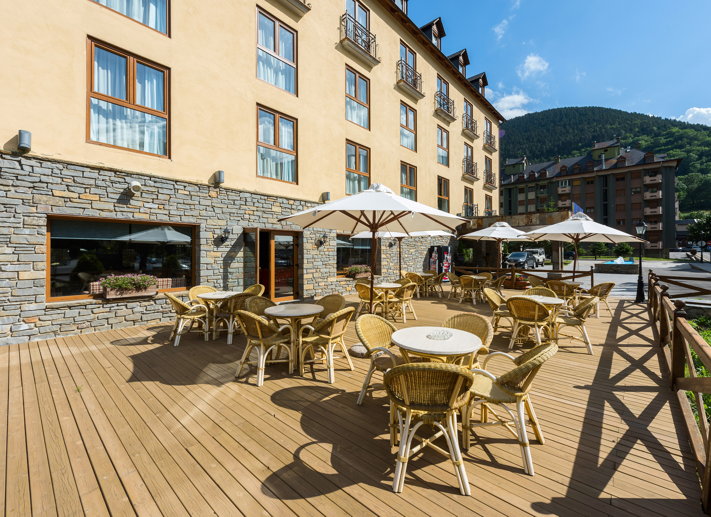 Terraza de hotel con mesas y sillas, parasoles, fachada de piedra y montaña.