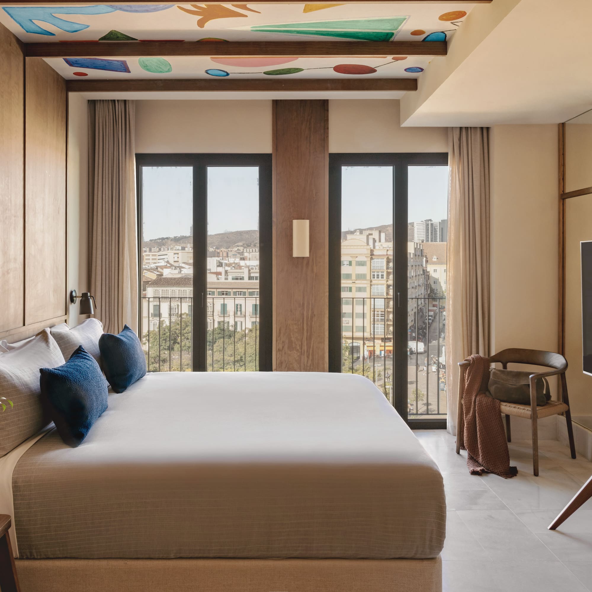 Modern hotel bedroom with king bed, city view, and colorful ceiling art.