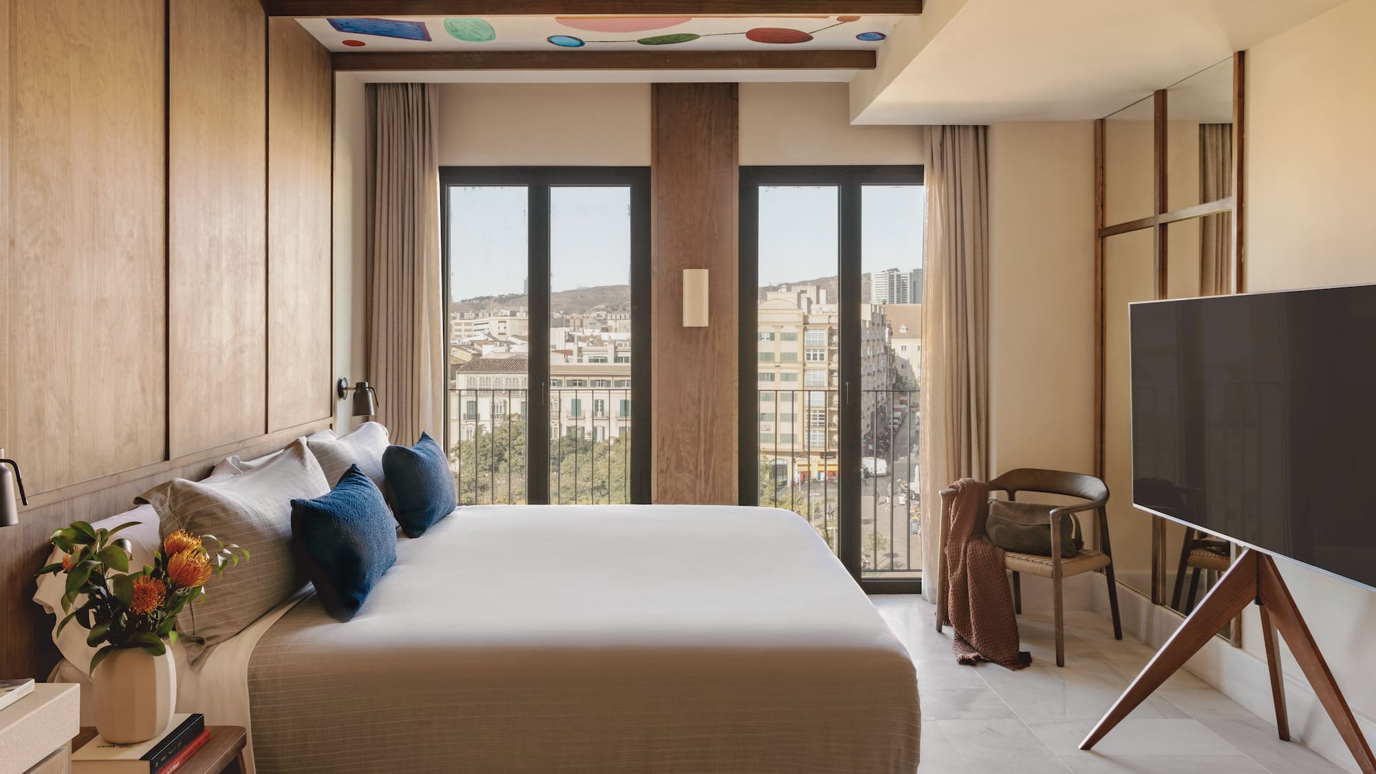 Modern hotel bedroom with king bed, city view, and colorful ceiling art.