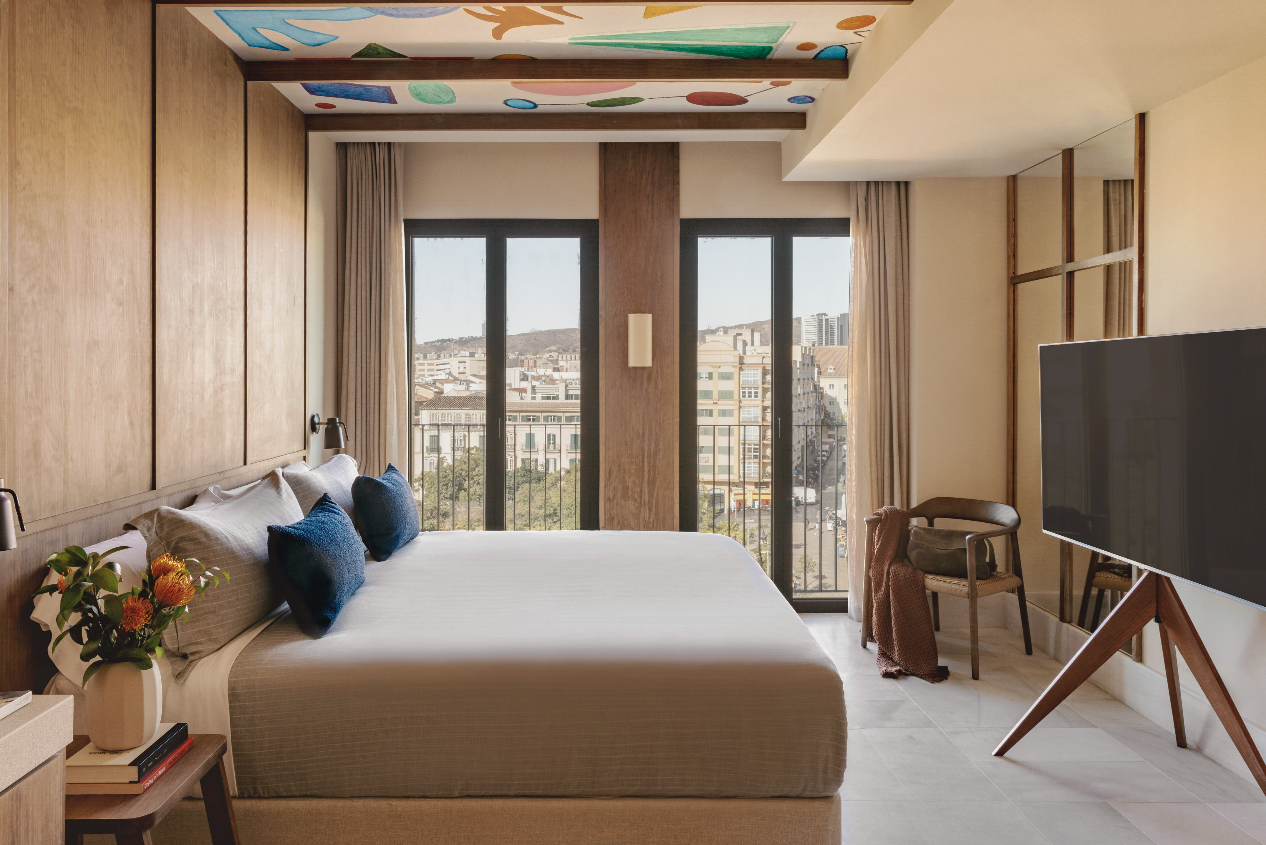 Modern hotel bedroom with king bed, city view, and colorful ceiling art.