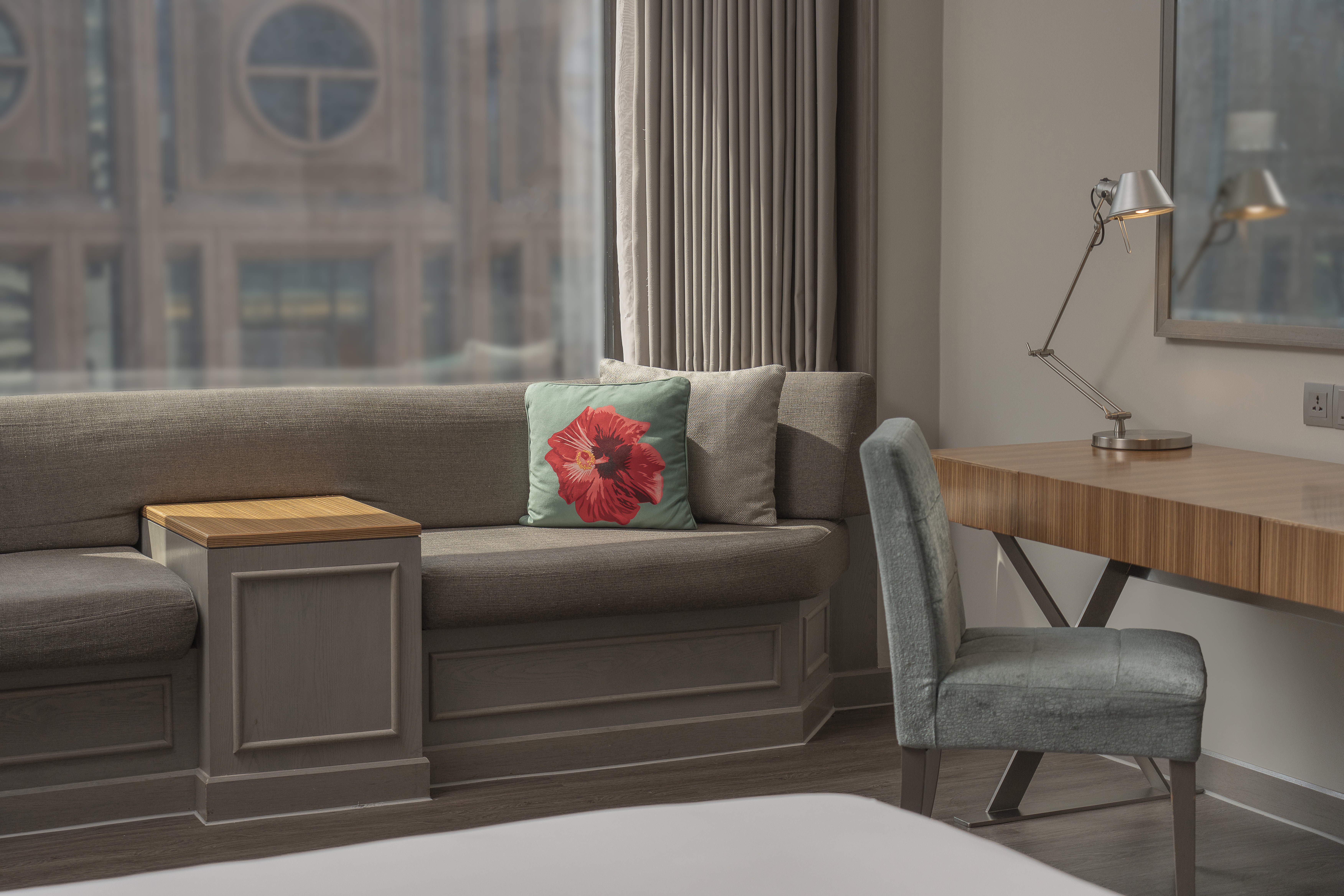 Hotel room interior: sofa with floral cushion, wooden desk, and task lamp.