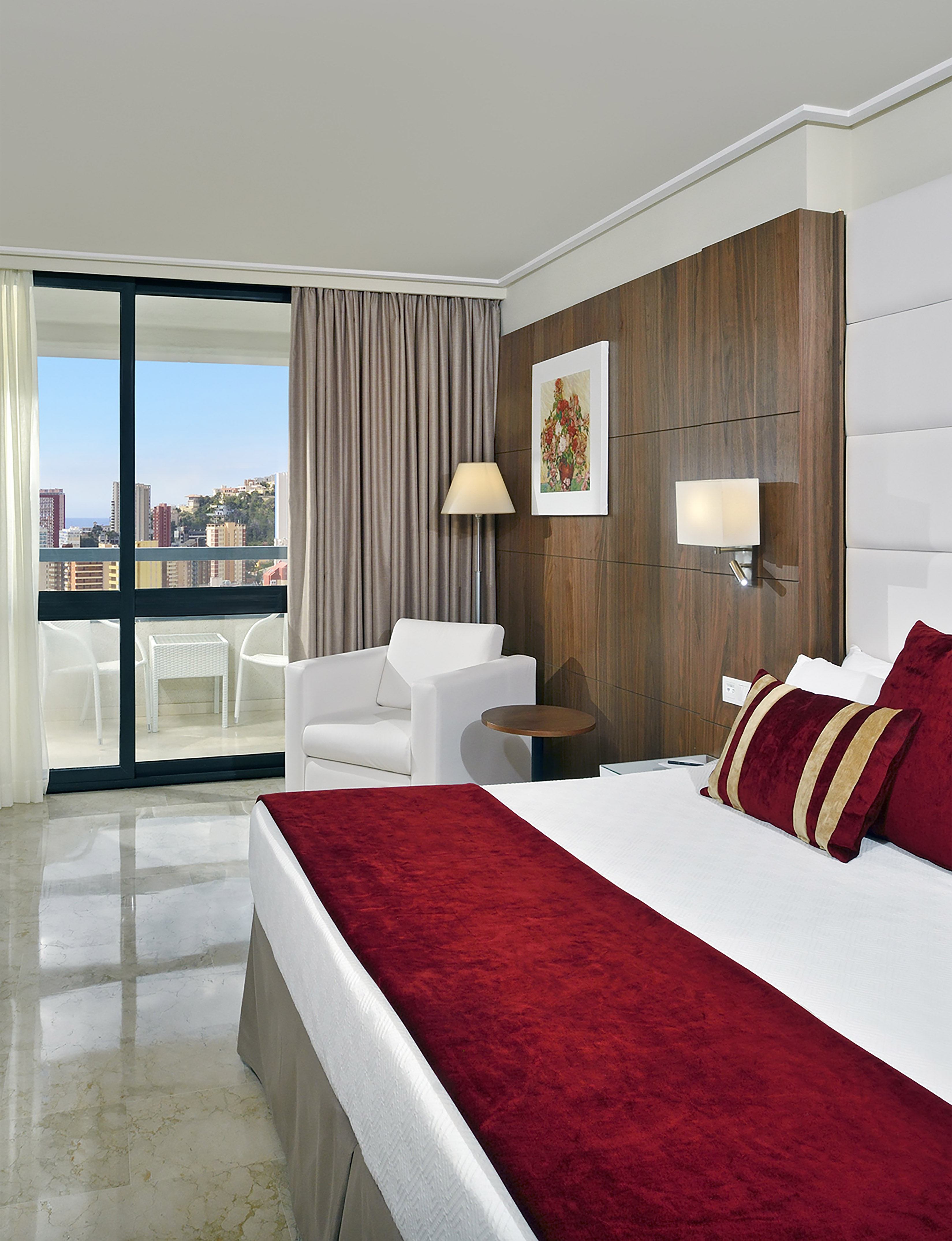Modern hotel room with red bedding, balcony city view, white armchair, wood paneling.