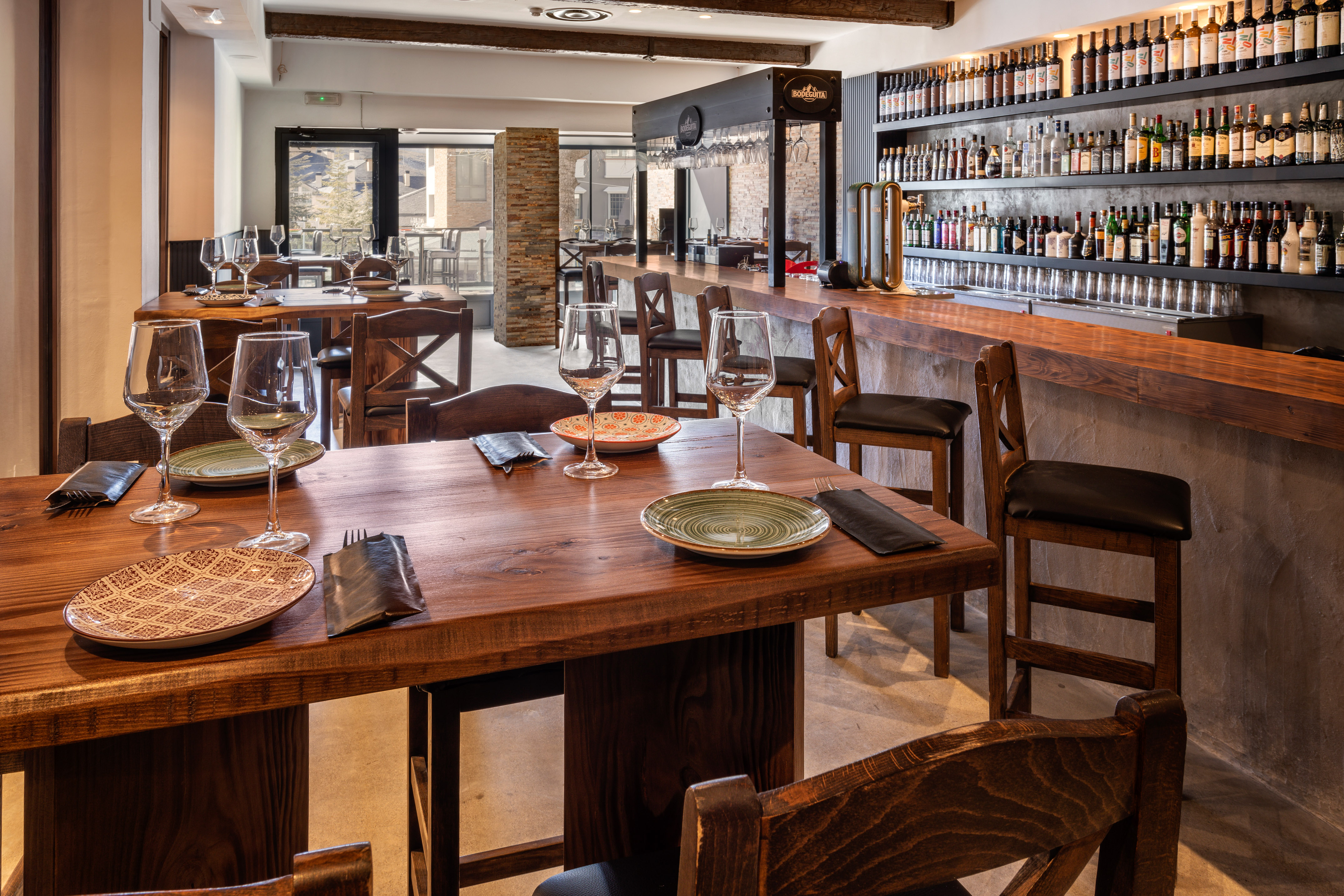 Rustic restaurant bar and dining area with dark wood tables, stools, and liquor shelves.