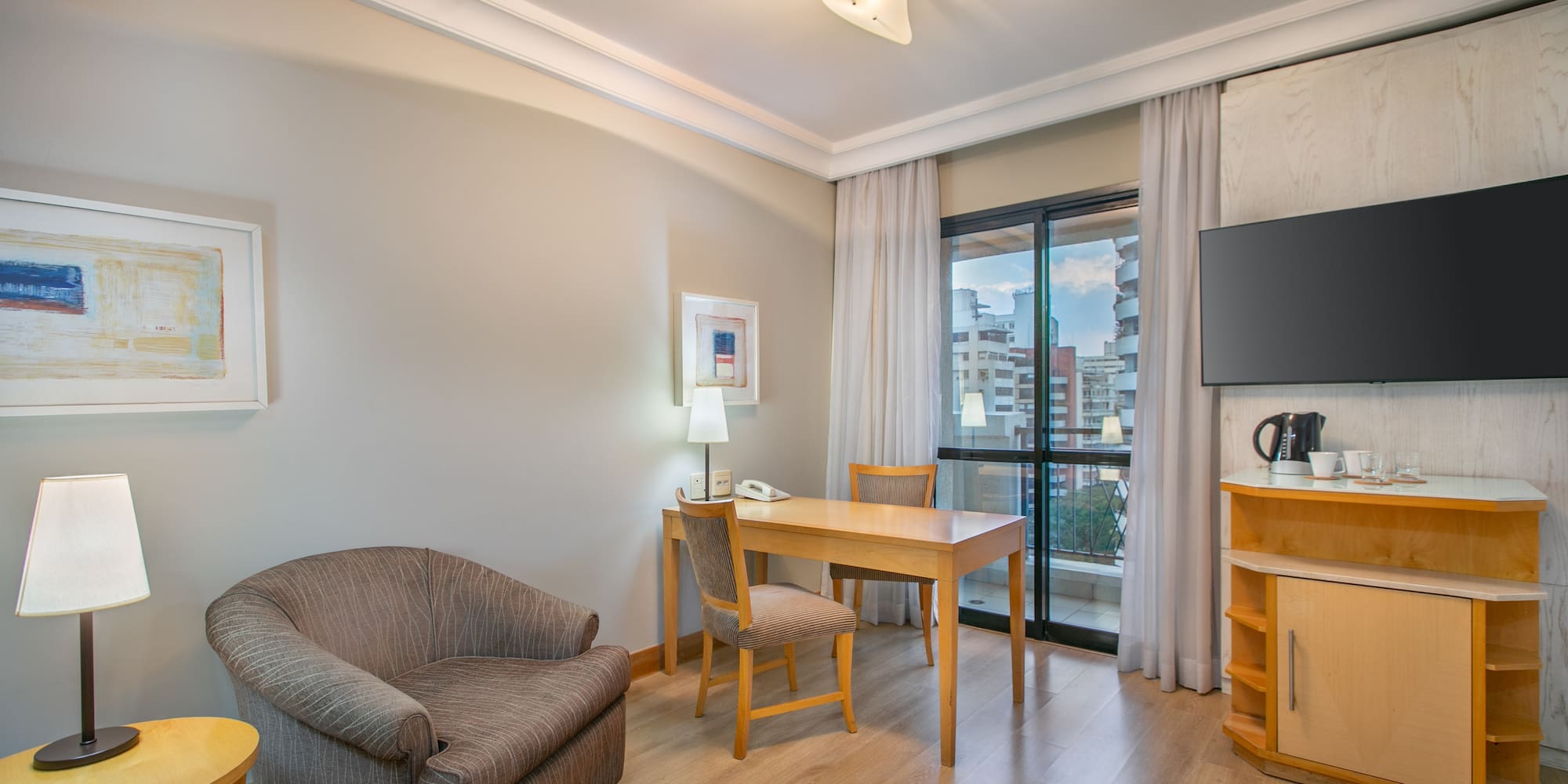Hotel suite living area with desk, TV, armchair, and city view.