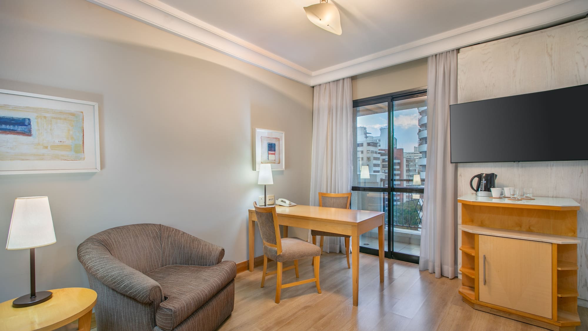 Hotel suite living area with desk, TV, armchair, and city view.