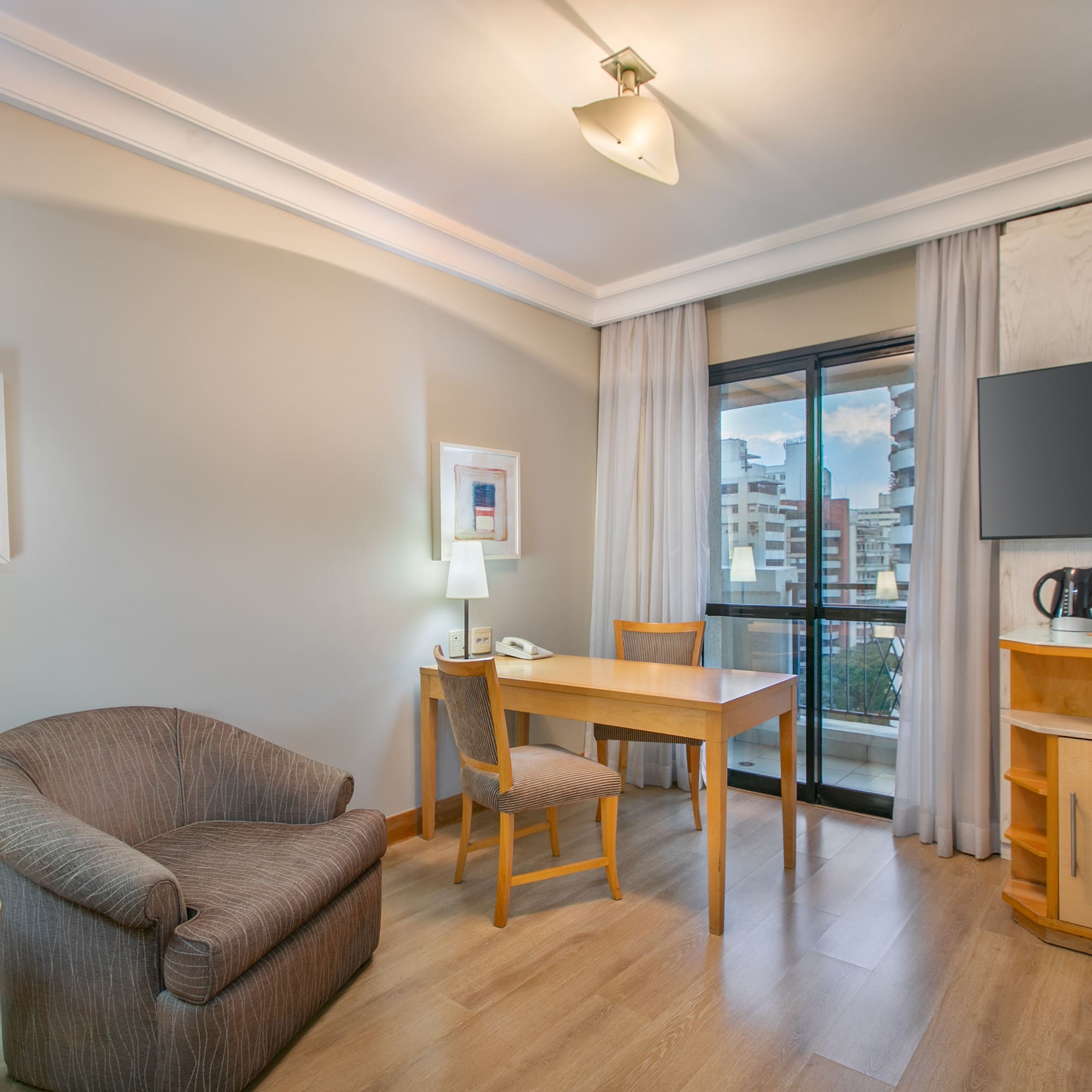 Hotel suite living area with desk, TV, armchair, and city view.