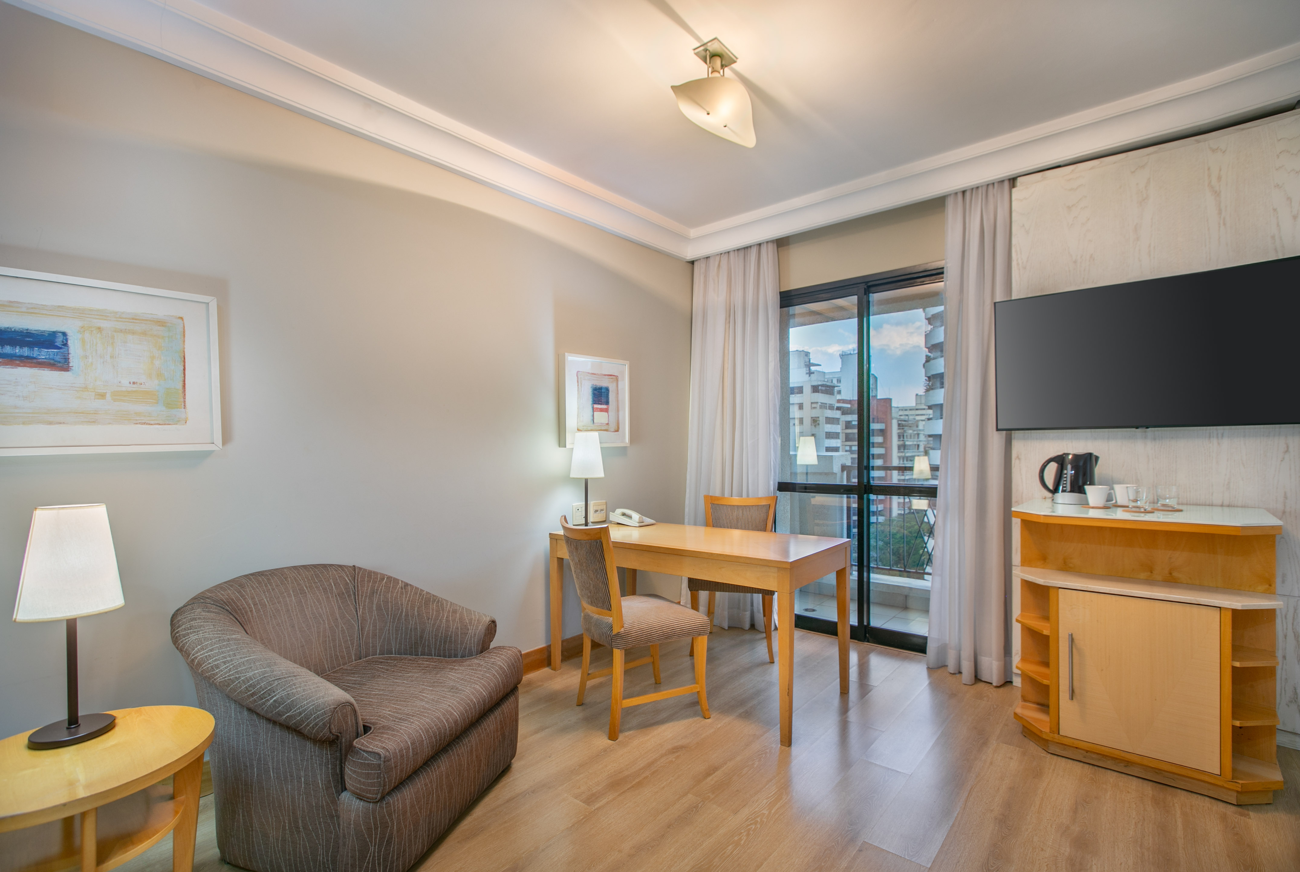 Hotel suite living area with desk, TV, armchair, and city view.