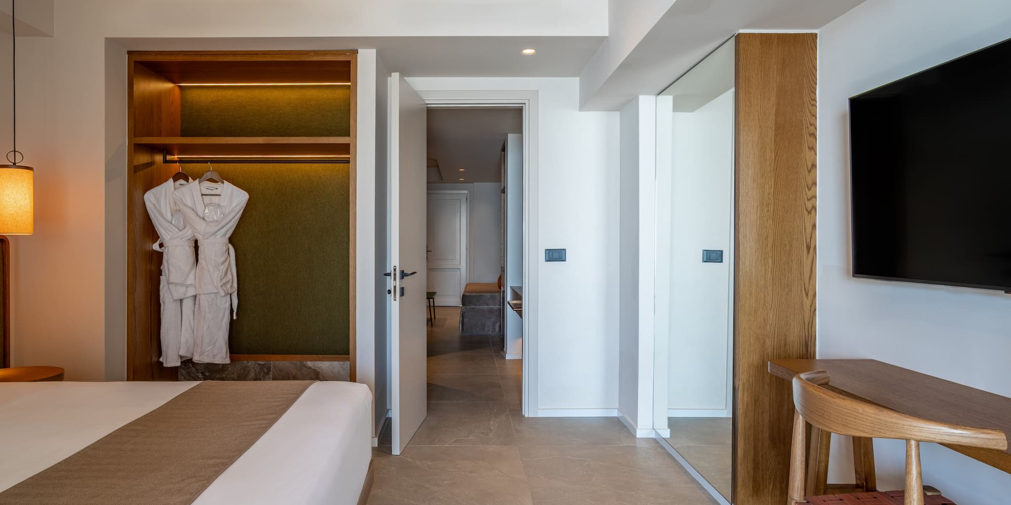 Modern hotel bedroom with open closet, bathrobes, large bed, and wall-mounted TV.