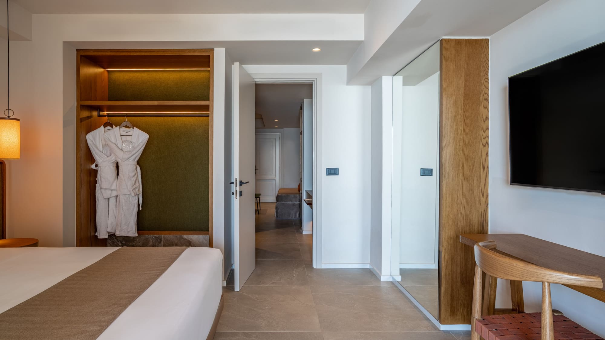 Modern hotel bedroom with open closet, bathrobes, large bed, and wall-mounted TV.
