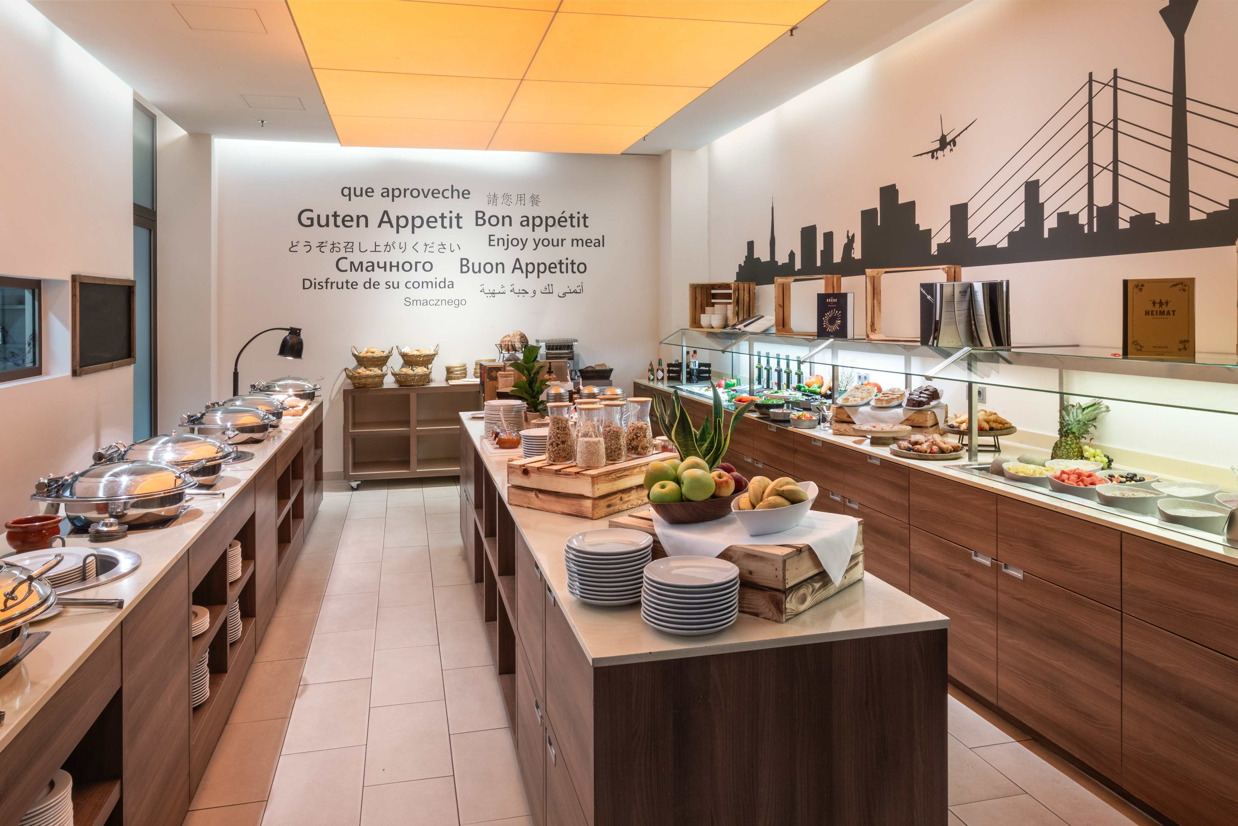 Hotel breakfast buffet with hot dishes, fresh fruit, pastries, and cityscape wall art.