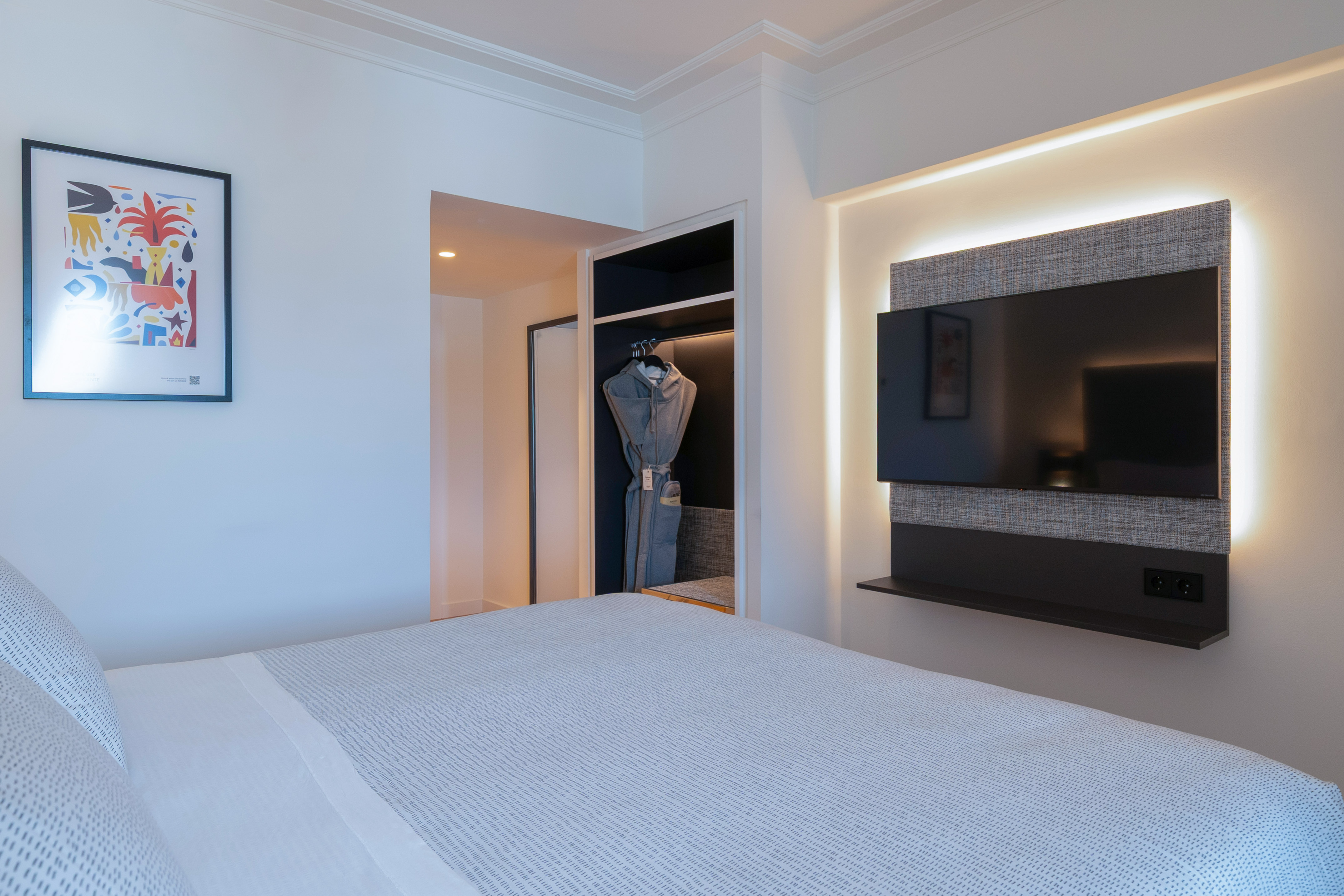 Habitación moderna de hotel: cama, TV retroiluminada, armario y cuadro abstracto.