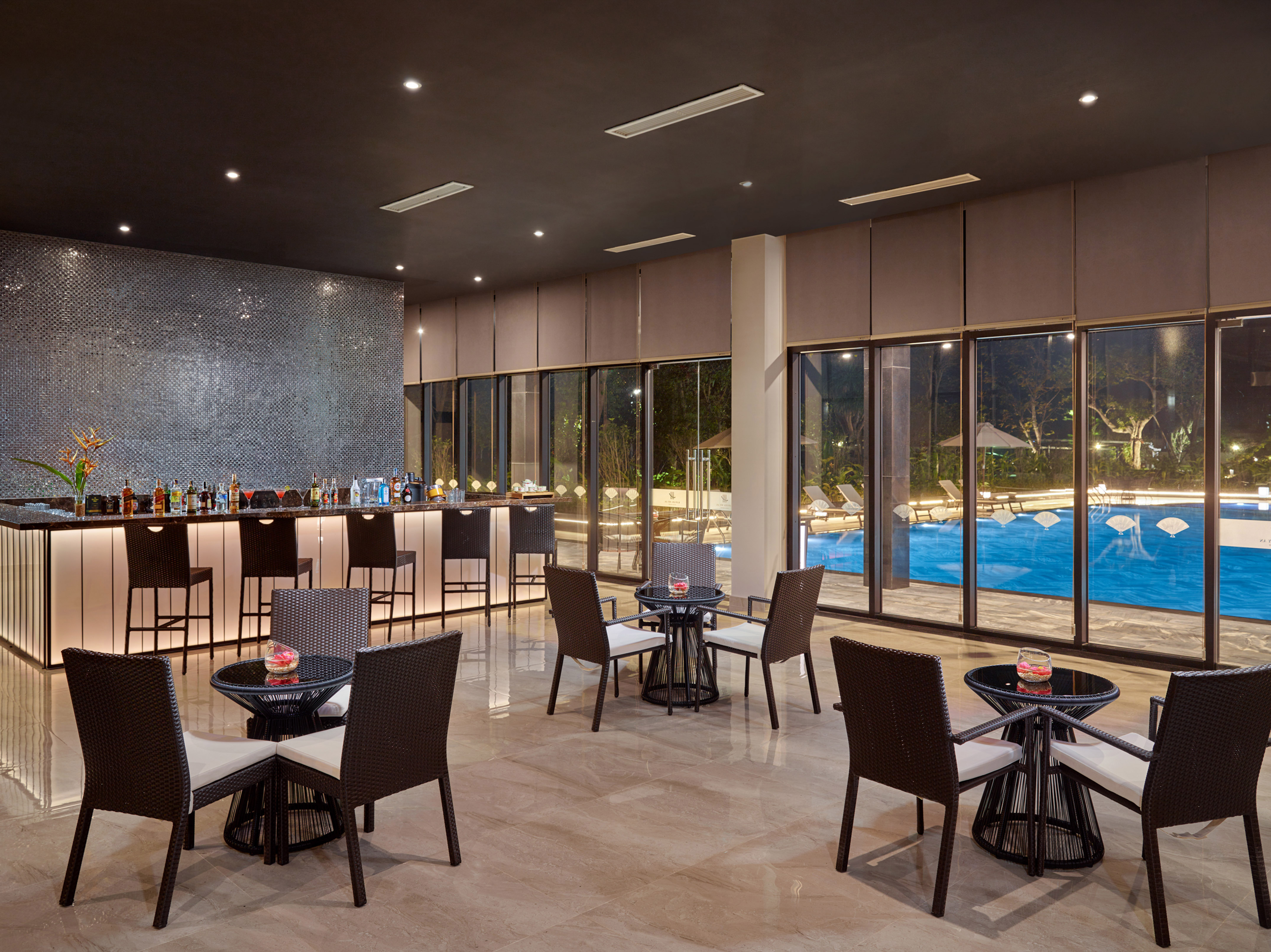 Modern hotel bar and lounge overlooking an illuminated swimming pool.