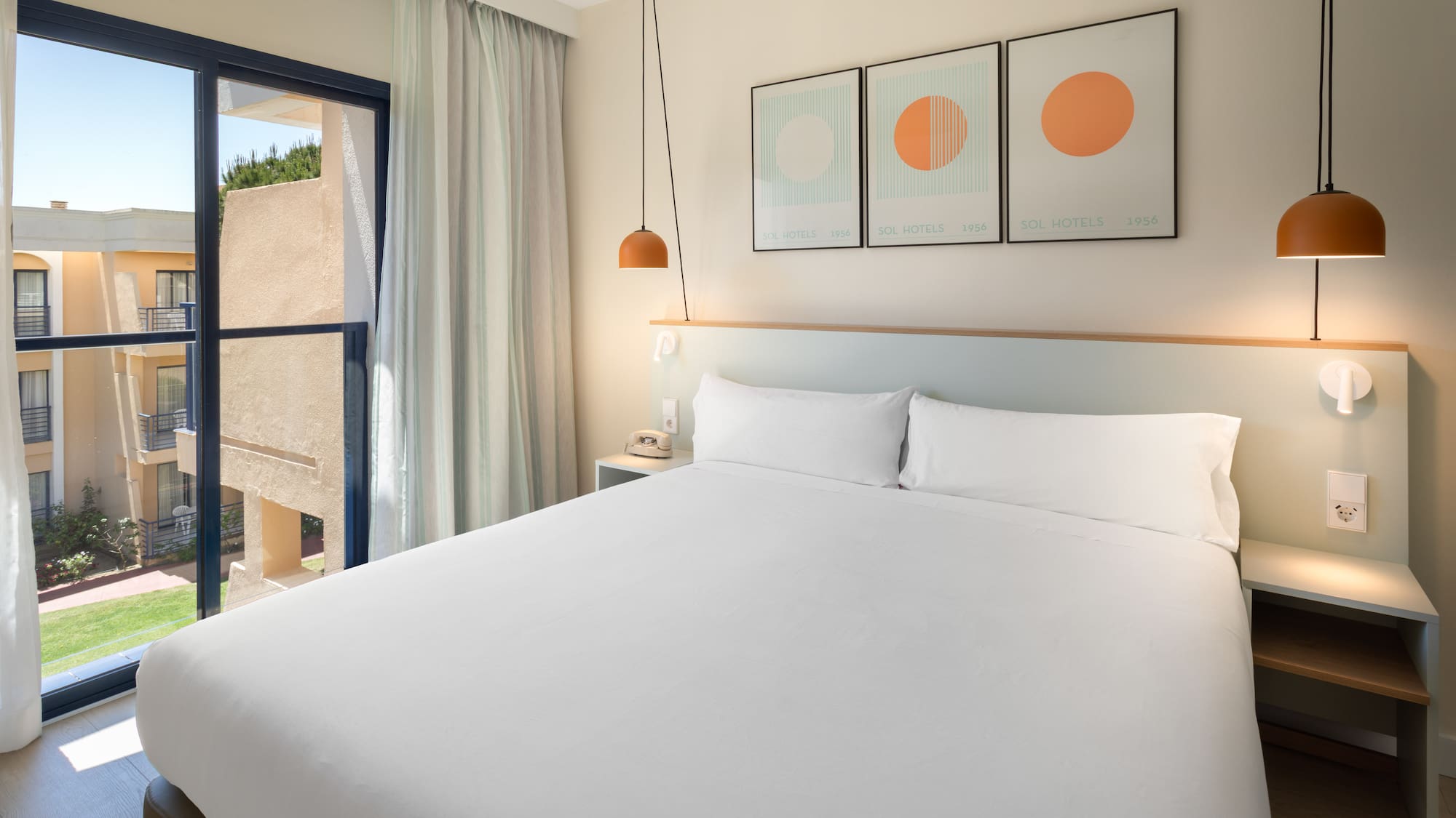 Modern hotel room with white bed, hanging lamps, and balcony view of resort.