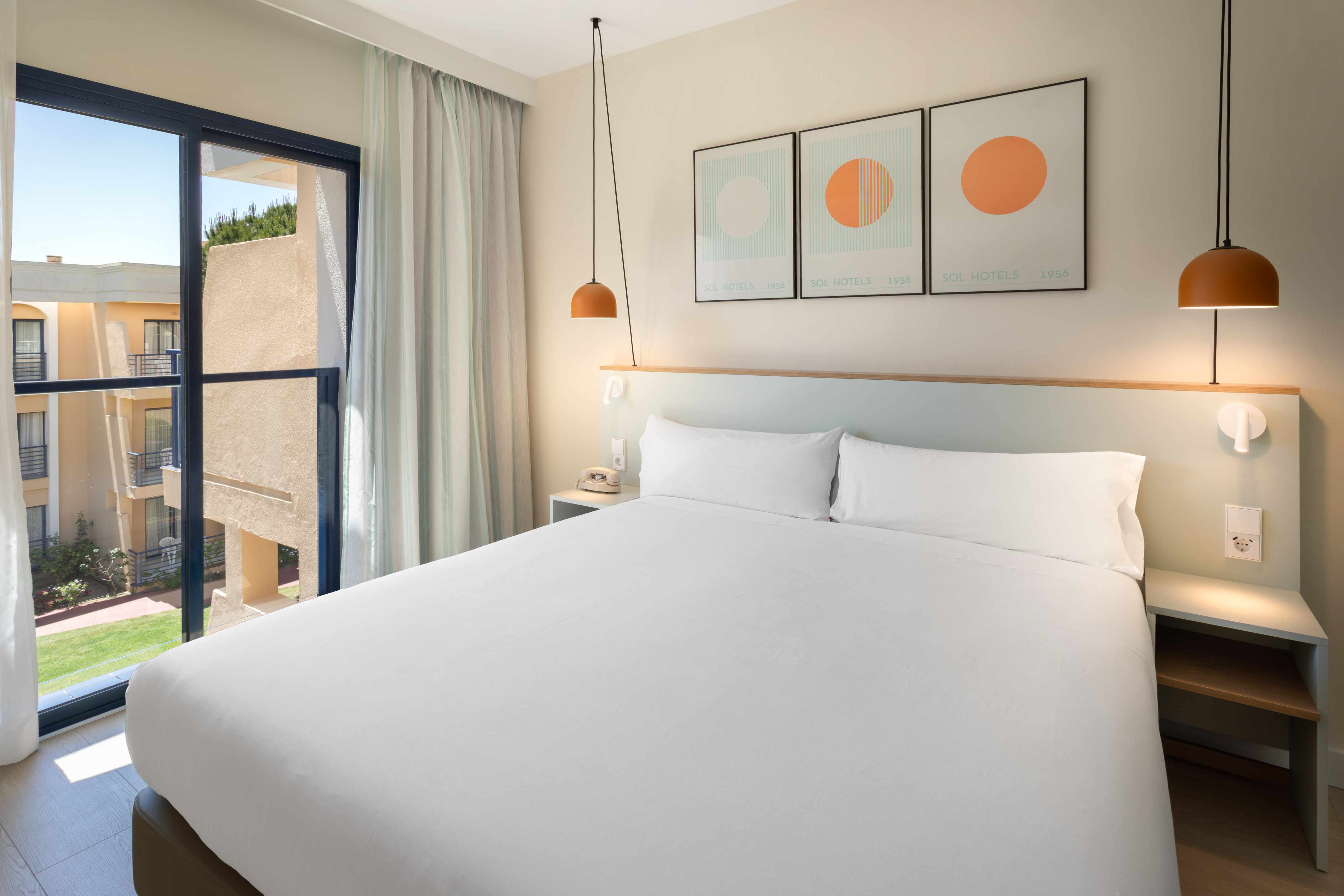 Modern hotel room with white bed, hanging lamps, and balcony view of resort.