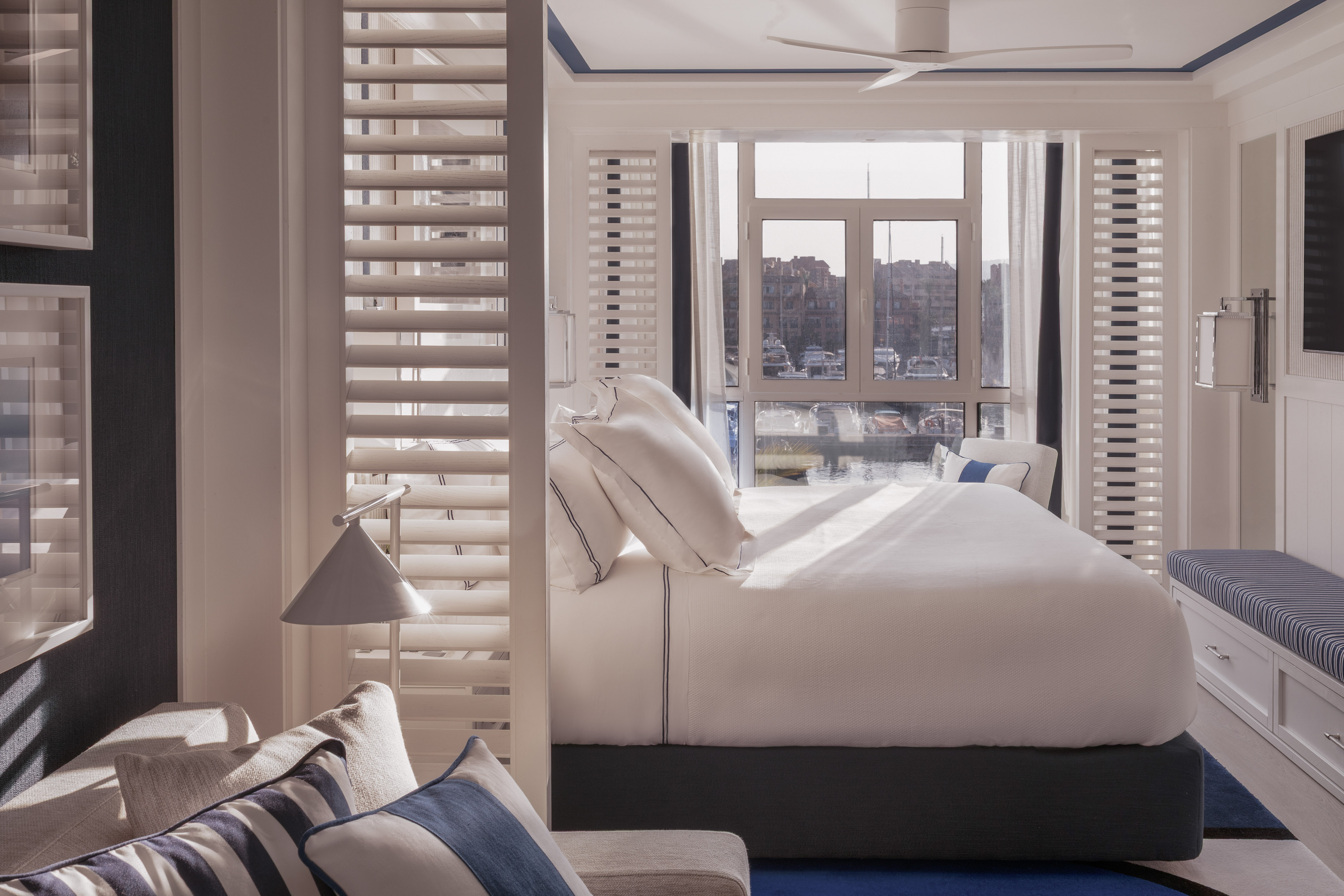 Coastal bedroom with white bed, marina view, wooden shutters, and blue accents.