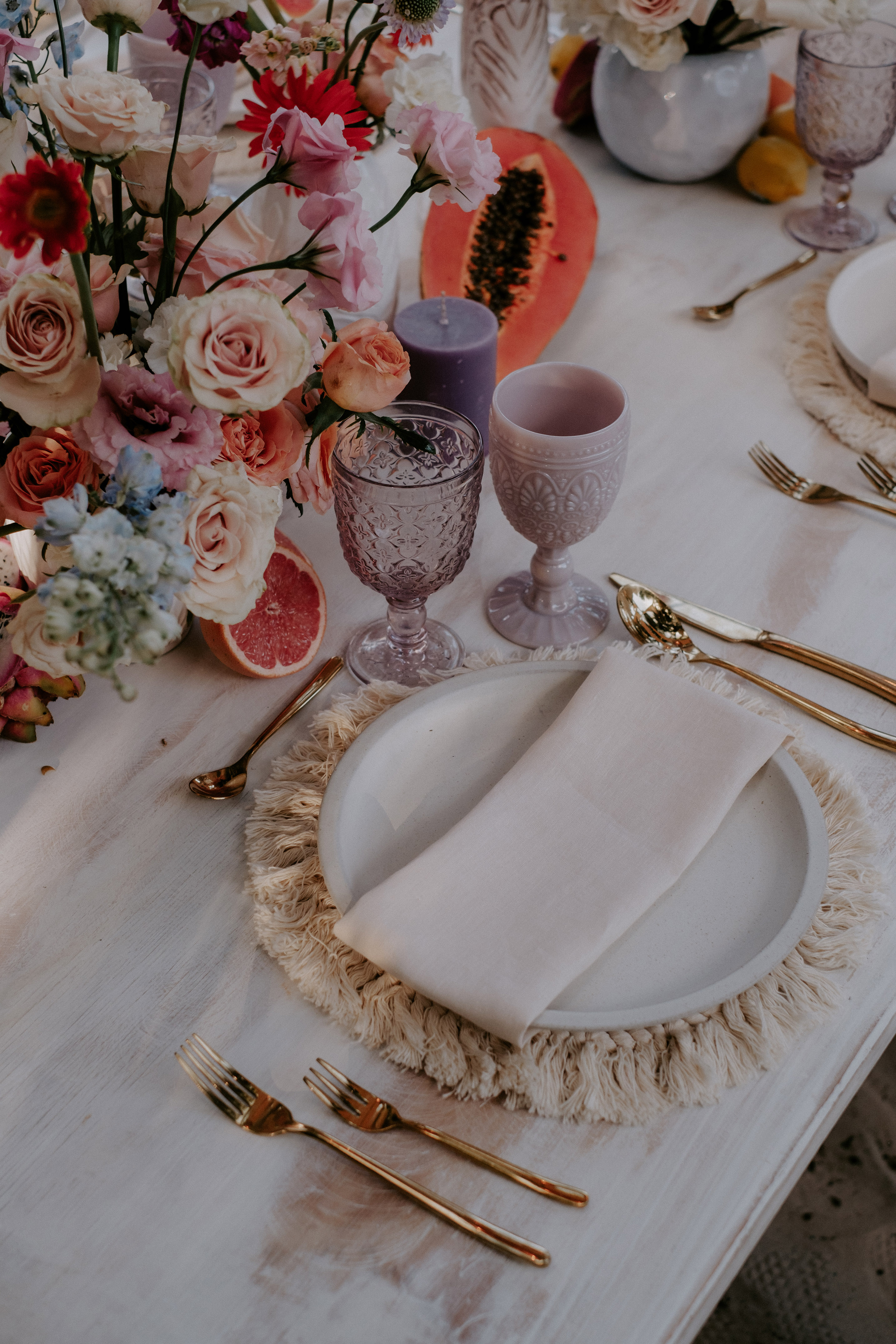 Boho table with roses, papaya, grapefruit, gold cutlery, and purple goblets.