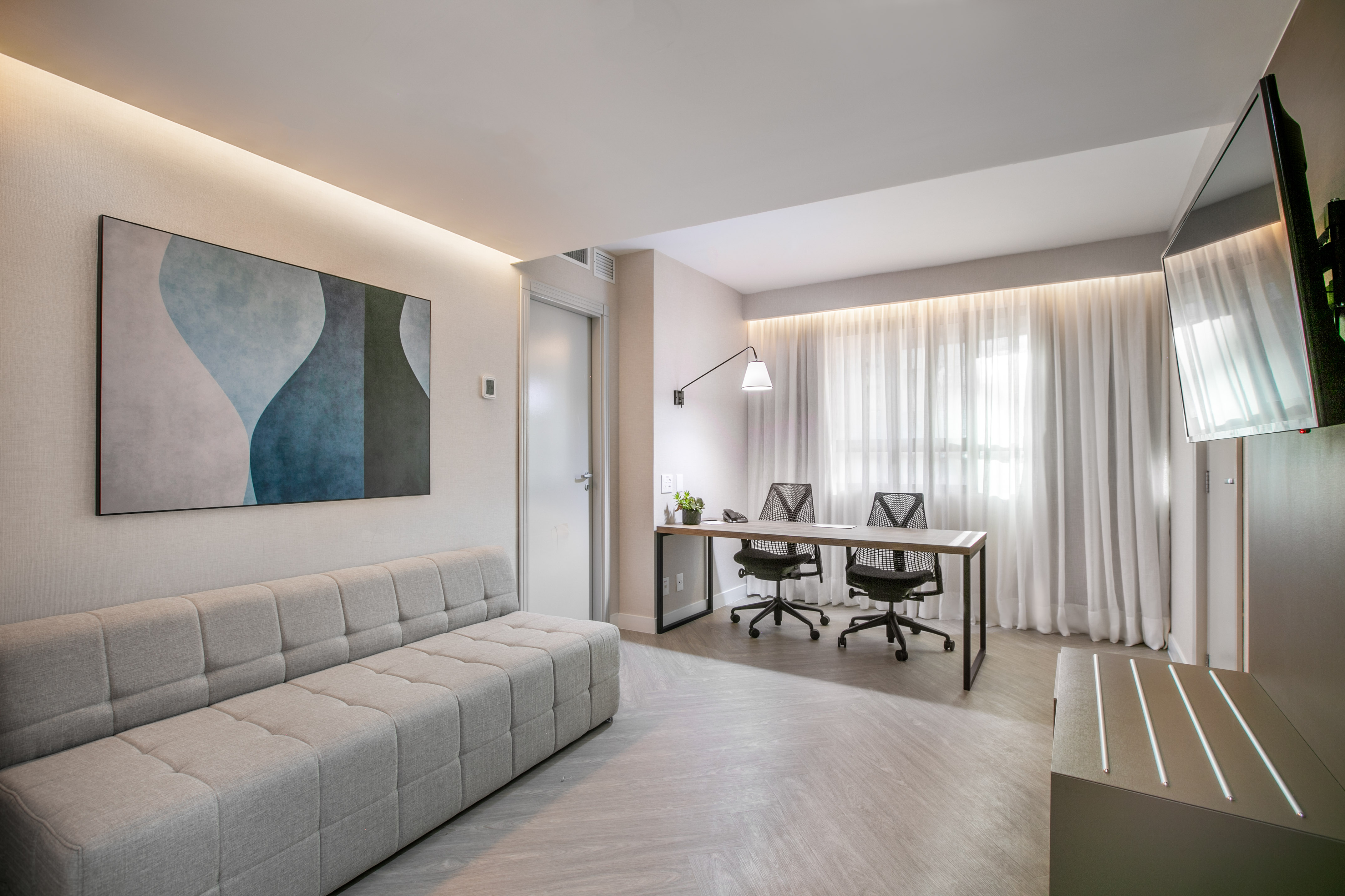 Modern suite with gray sofa, dual work desk, abstract art, and sheer curtains.