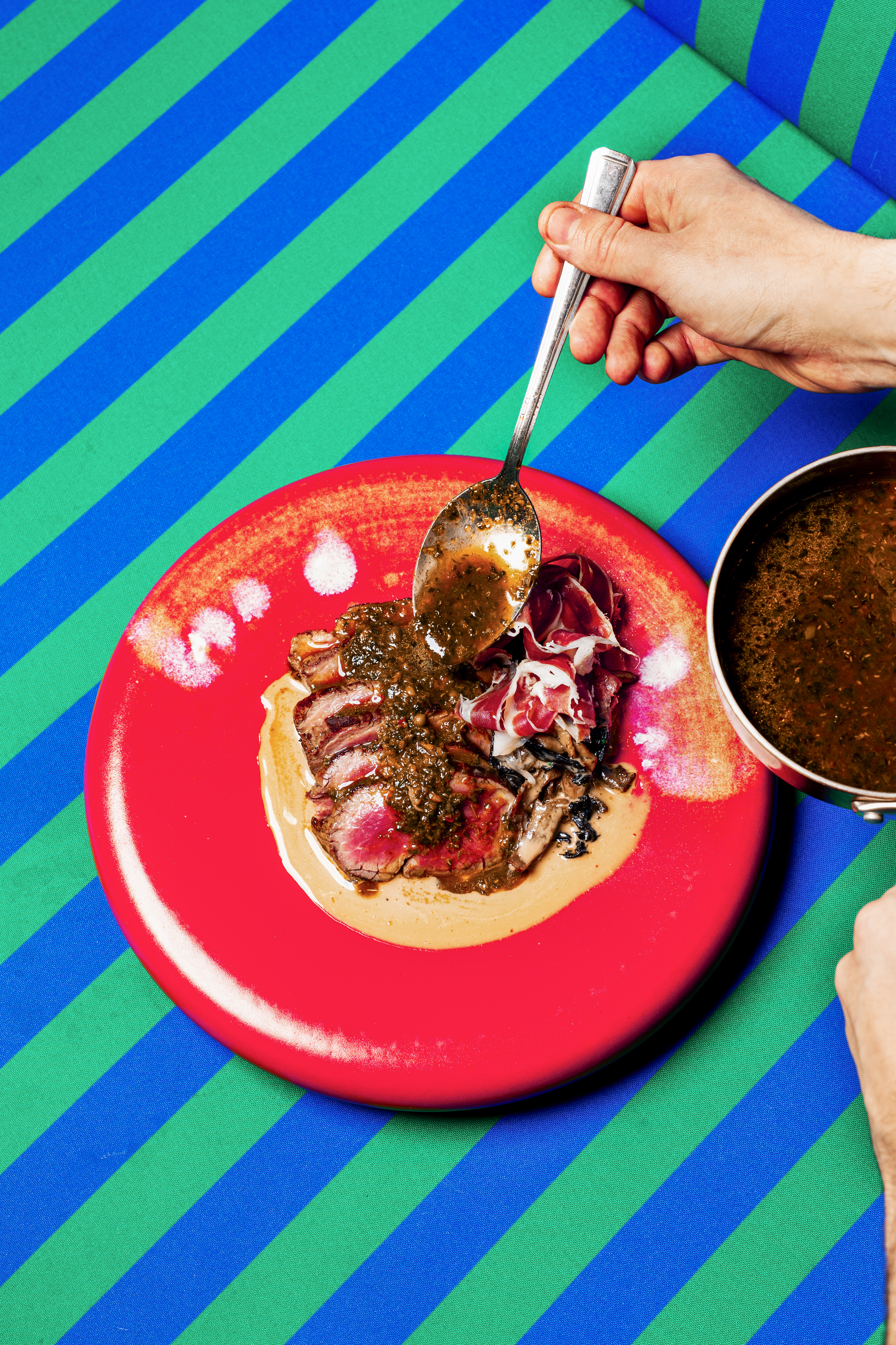 Hand serving sliced steak with sauce, prosciutto, mushrooms on a red plate.
