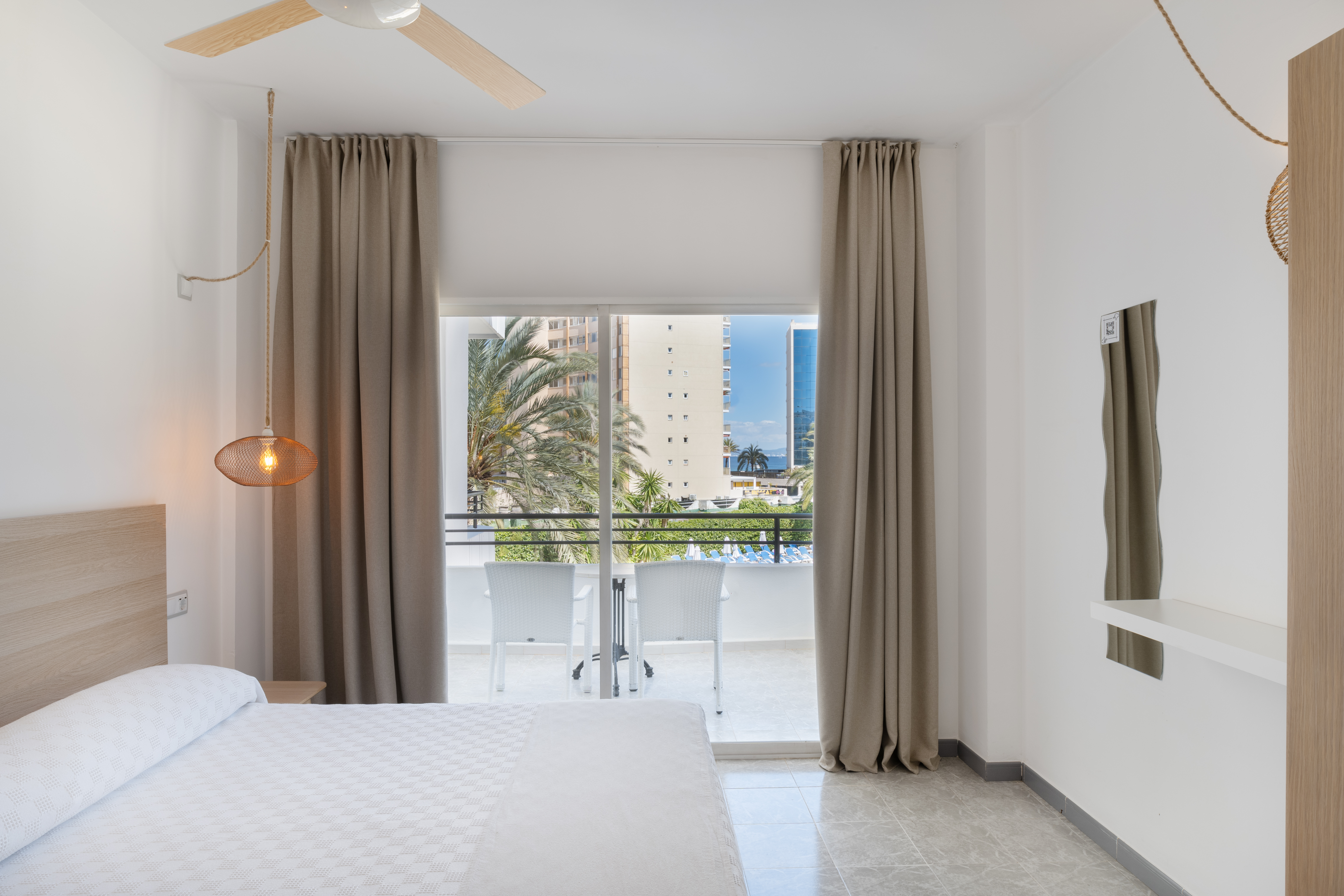 Bright hotel bedroom with white bed, private balcony, and tropical city view.