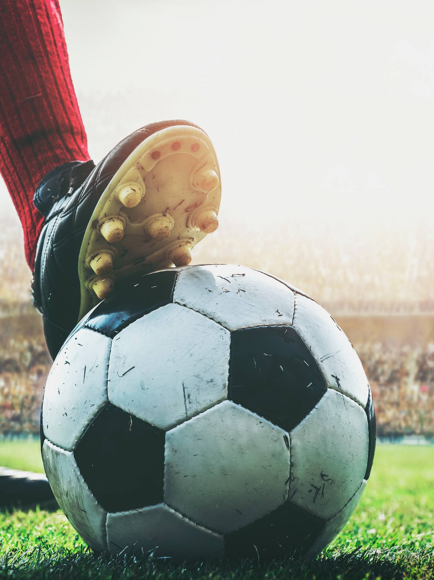 Soccer player's cleat on ball, red sock, green field, stadium crowd.