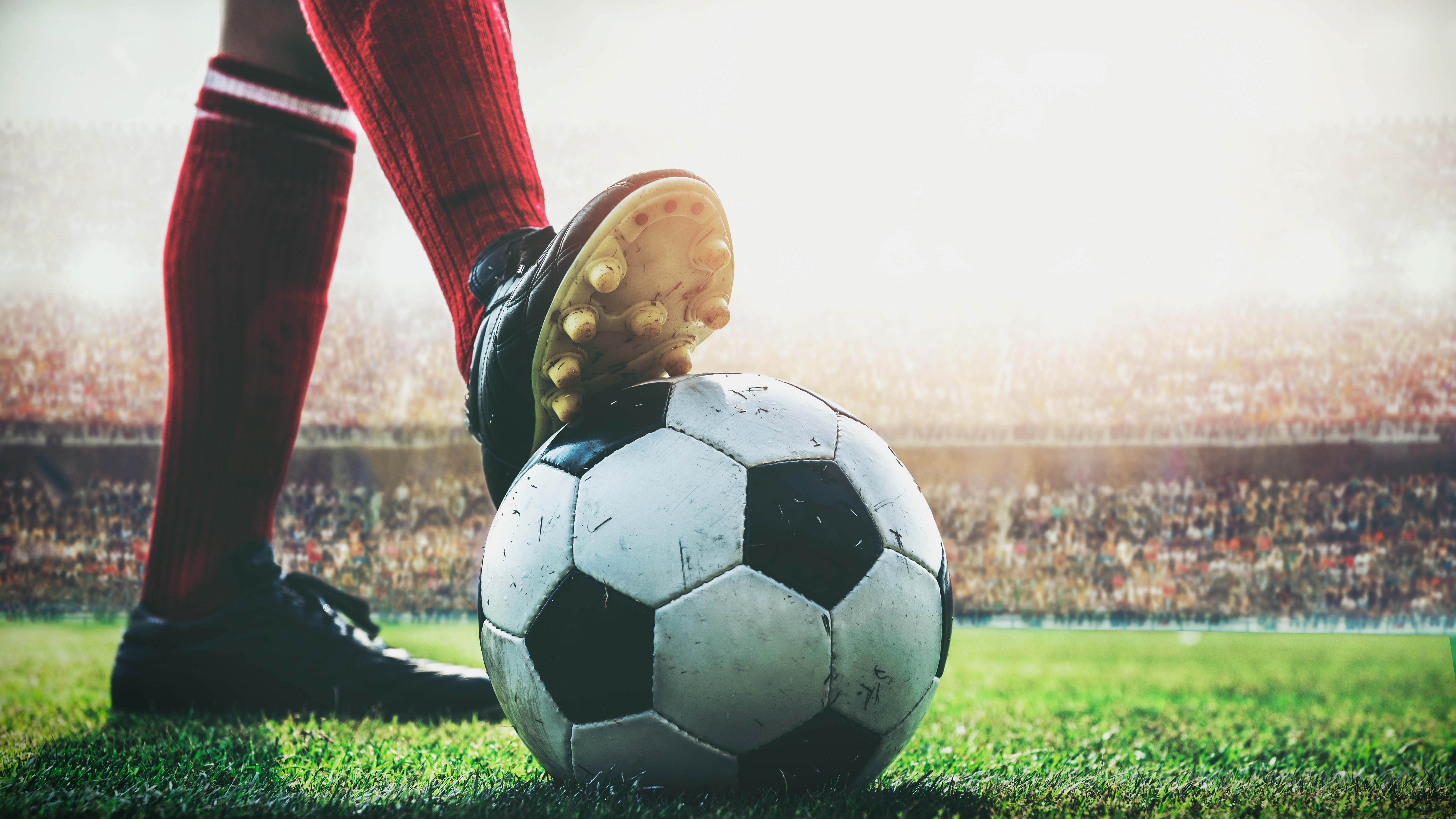 Soccer player's cleat on ball, red sock, green field, stadium crowd.
