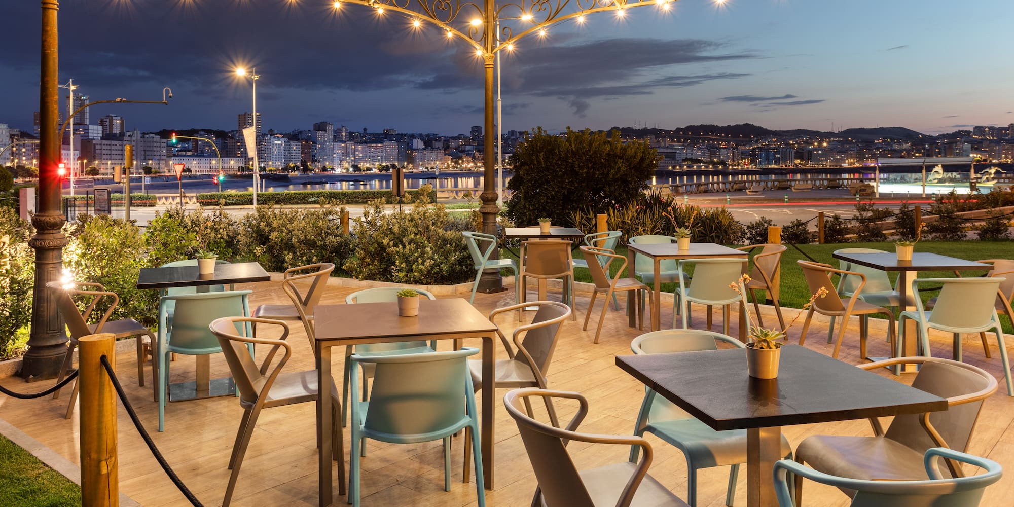 Evening cafe patio with modern chairs, string lights, city waterfront view.
