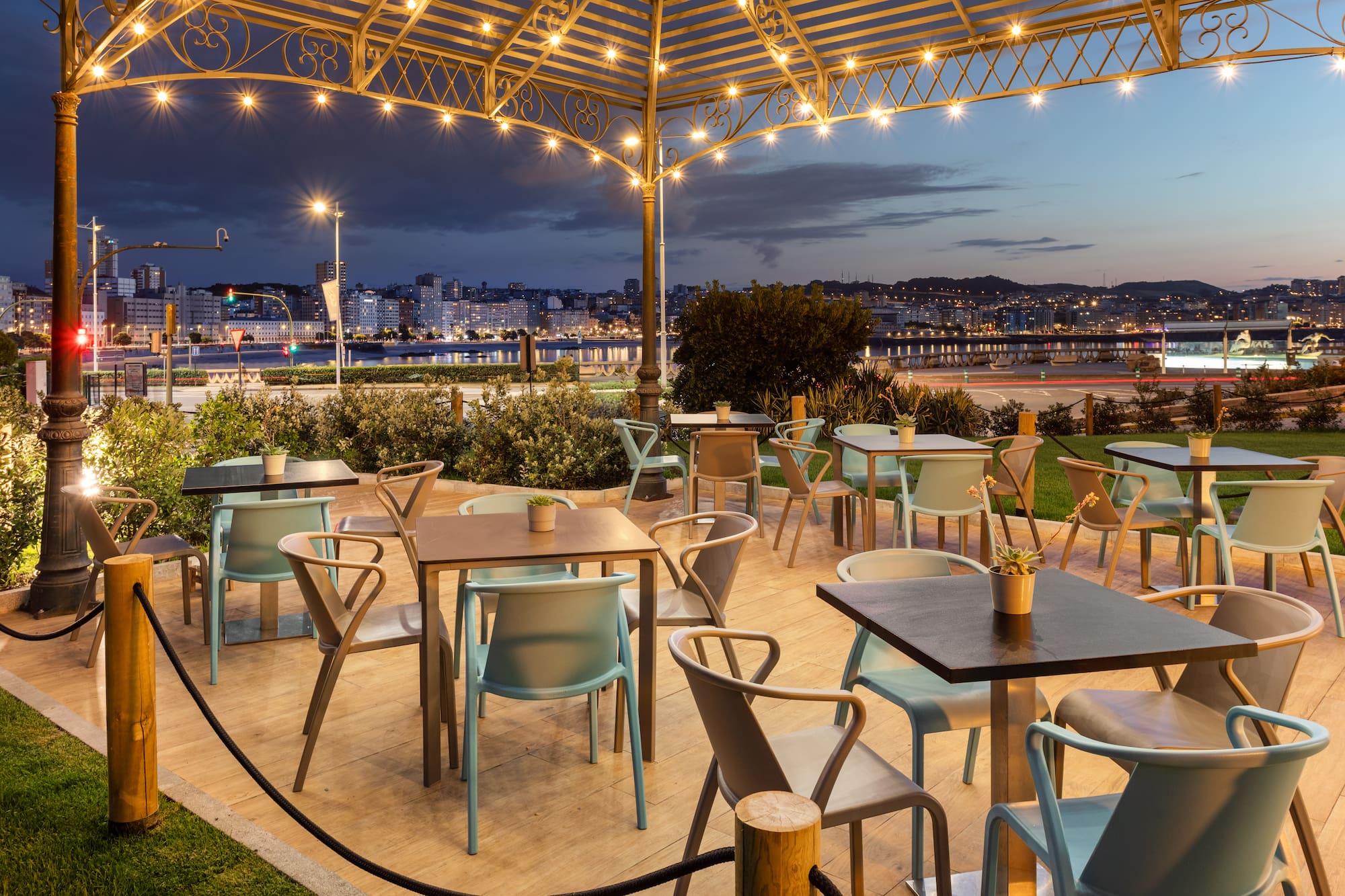 Evening cafe patio with modern chairs, string lights, city waterfront view.