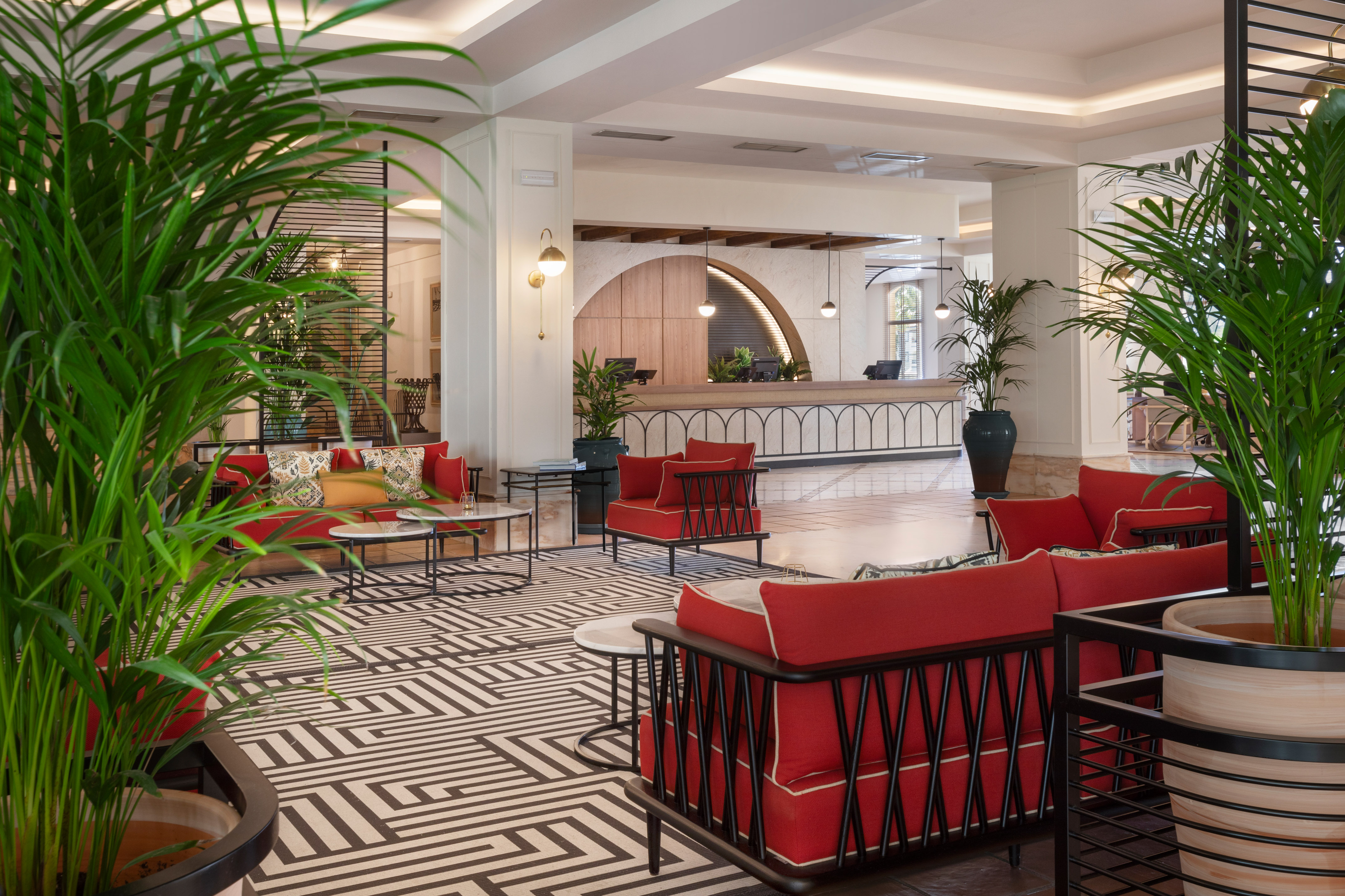Elegant hotel lobby with red sofas, green palm plants, and reception area.
