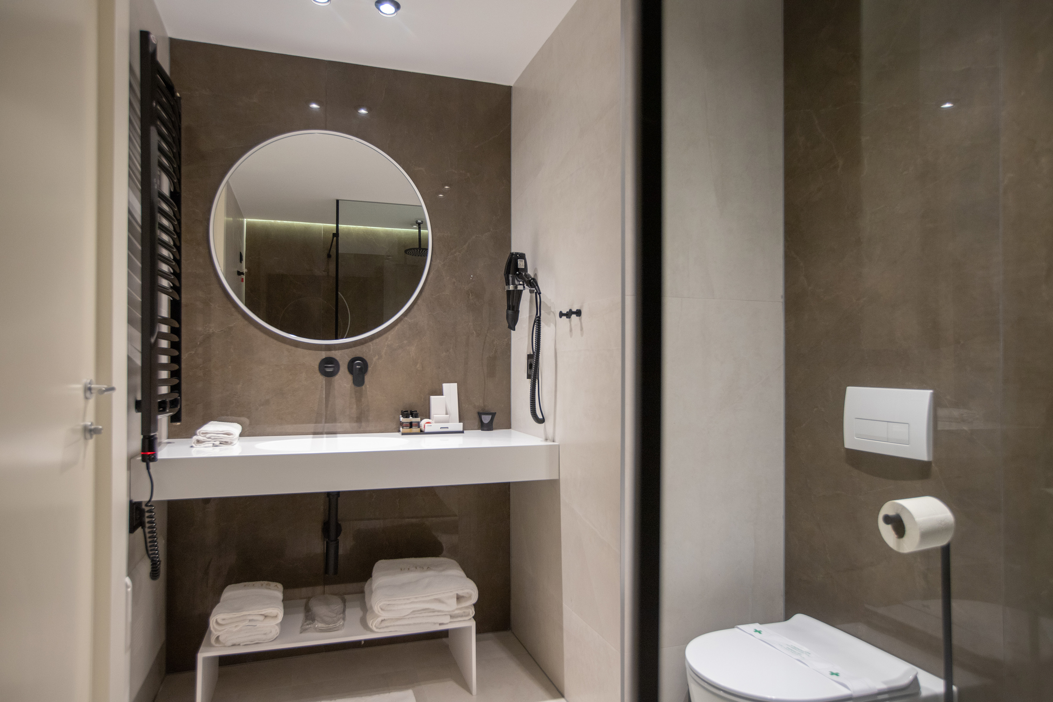 Modern hotel bathroom with round mirror, white vanity, and tiled walls.