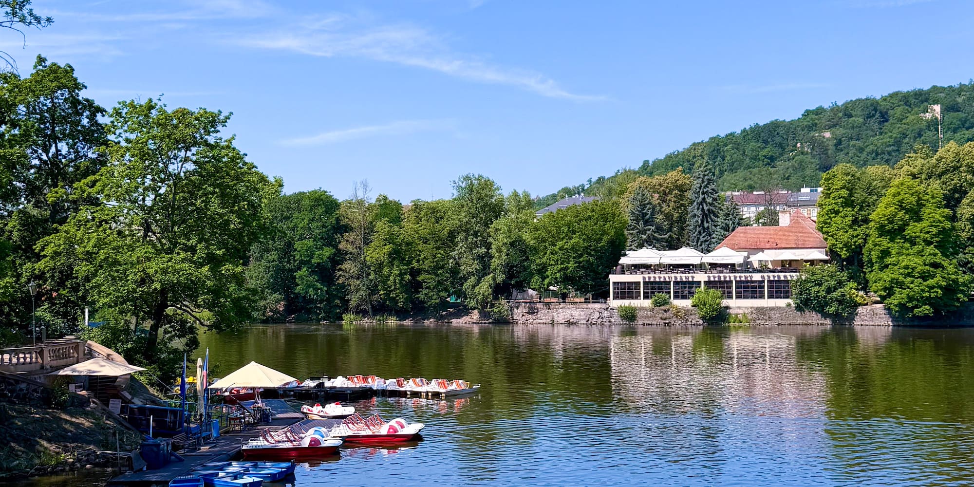 Colorful pedal boats on a lake with a waterside restaurant and green trees.