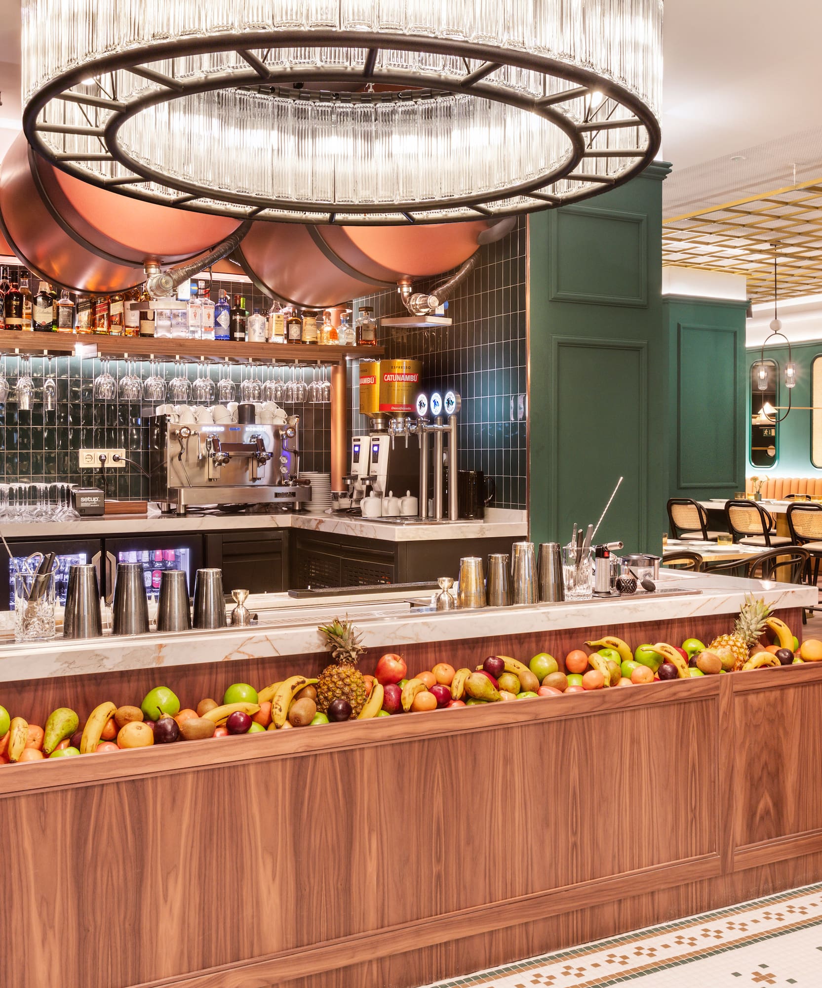 Modern bar with fresh fruit counter, green walls, copper accents, and chandelier.