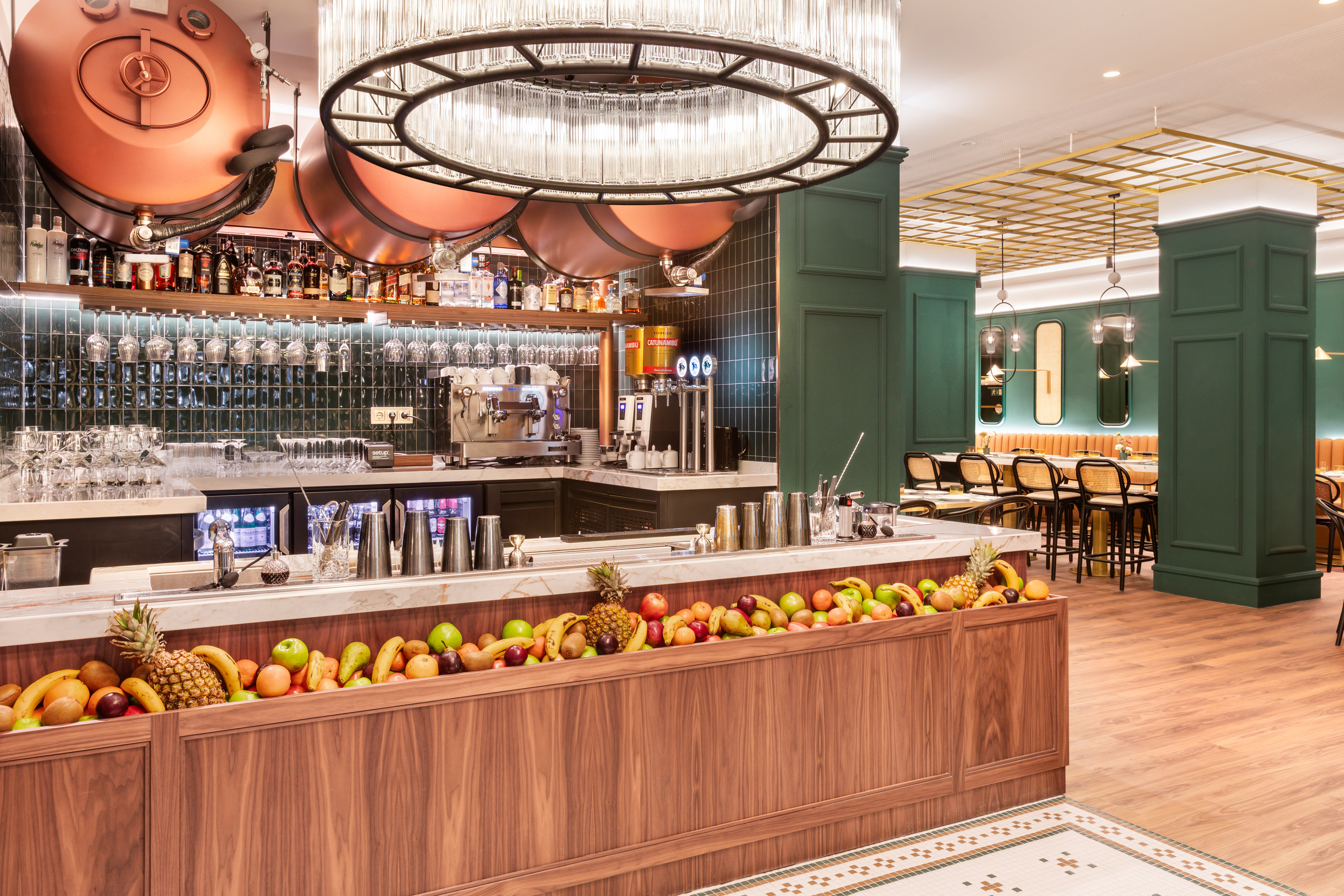 Modern bar with fresh fruit counter, green walls, copper accents, and chandelier.