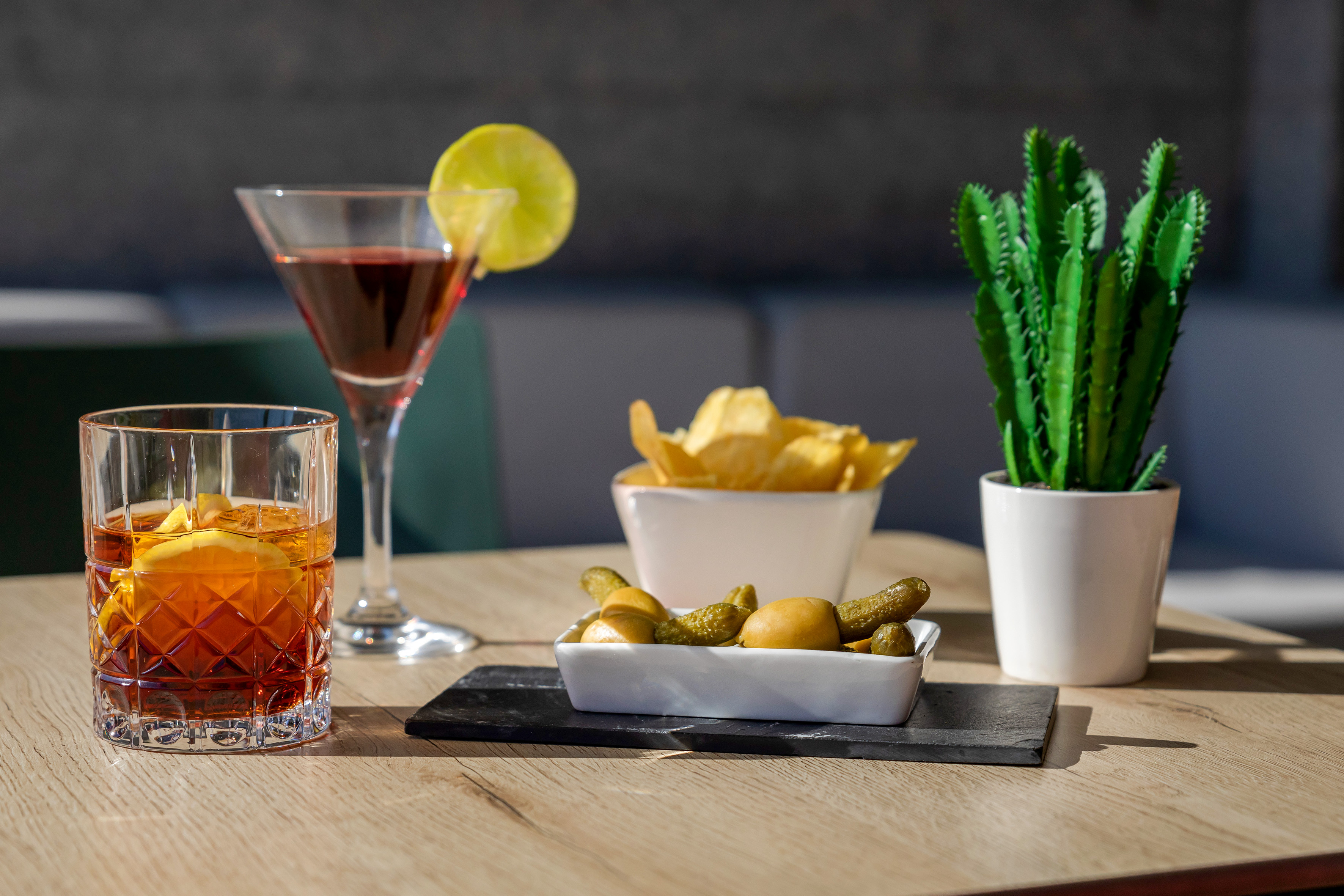 Cocktails, chips, pickles, and olives on a sunny wooden table.
