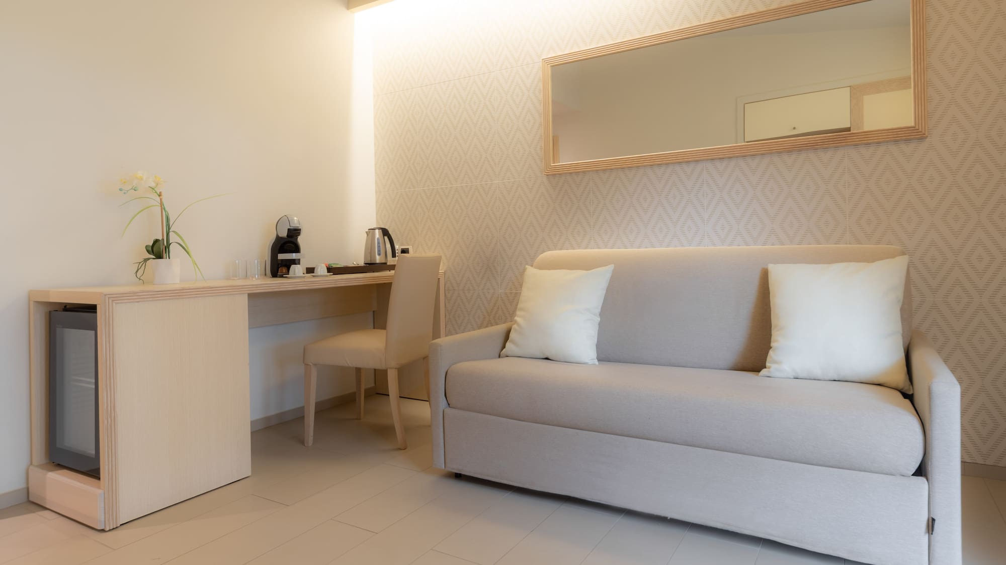 Modern hotel room with sofa, desk, mini-fridge, Nespresso machine, and wall mirror.