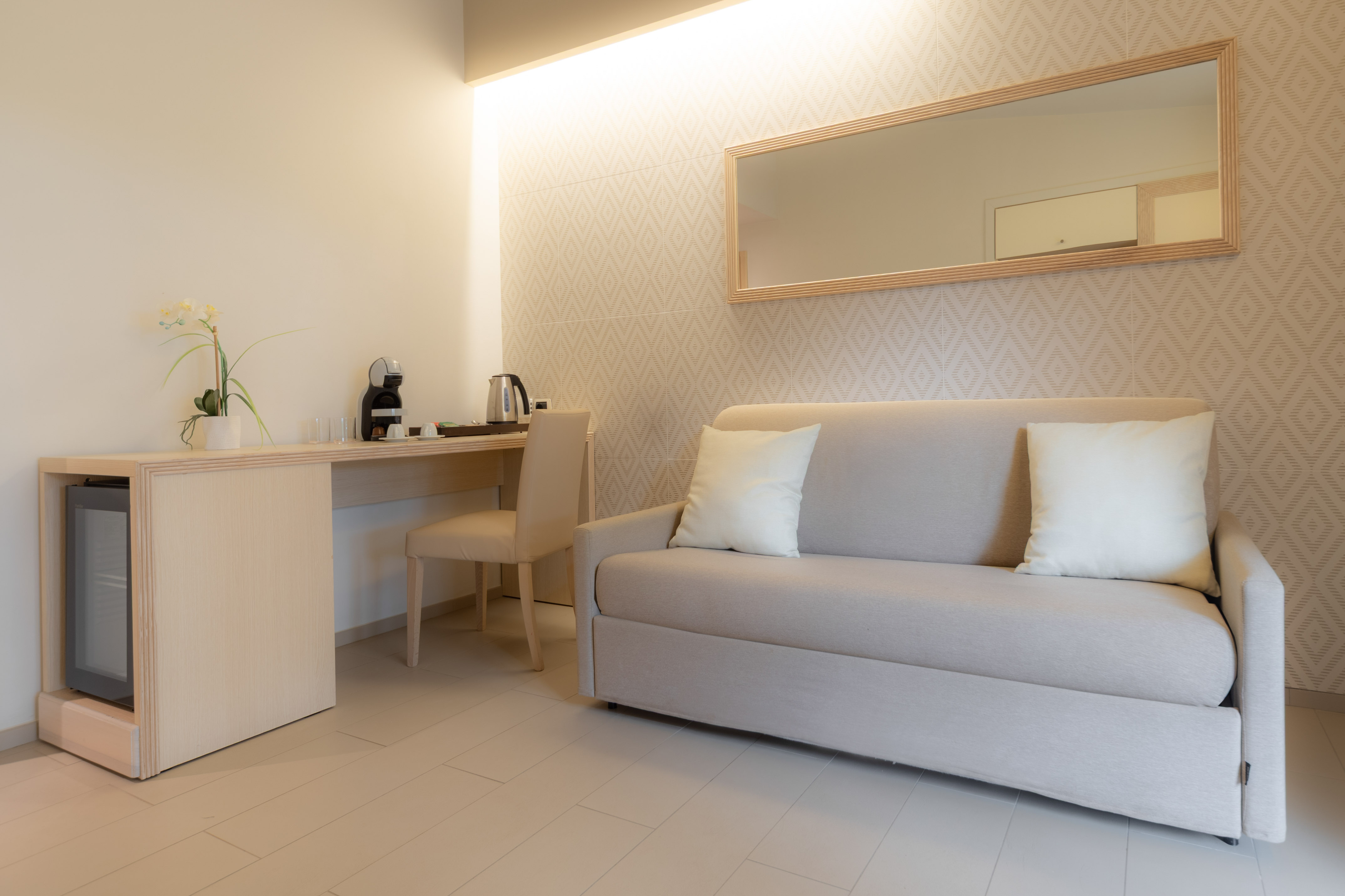 Modern hotel room with sofa, desk, mini-fridge, Nespresso machine, and wall mirror.