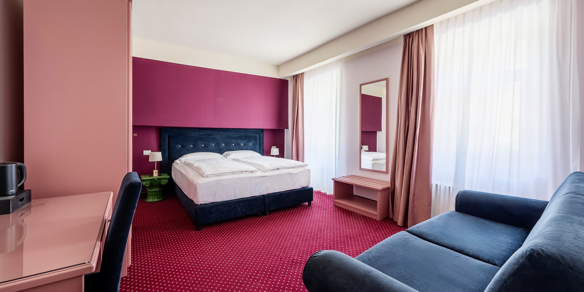 Vibrant hotel room: blue bed, red carpet, pink/magenta accent wall, blue sofa.