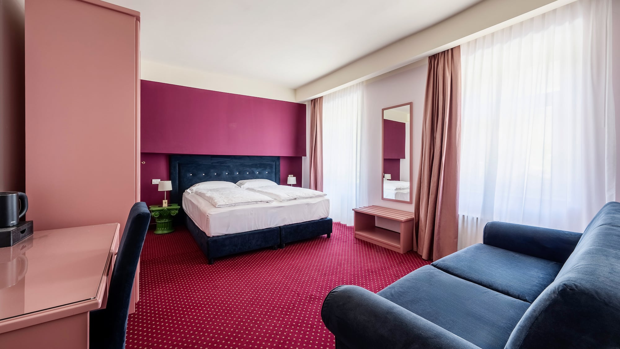 Hotel bedroom with red carpet, blue bed, magenta accent wall, pink desk, and blue sofa.