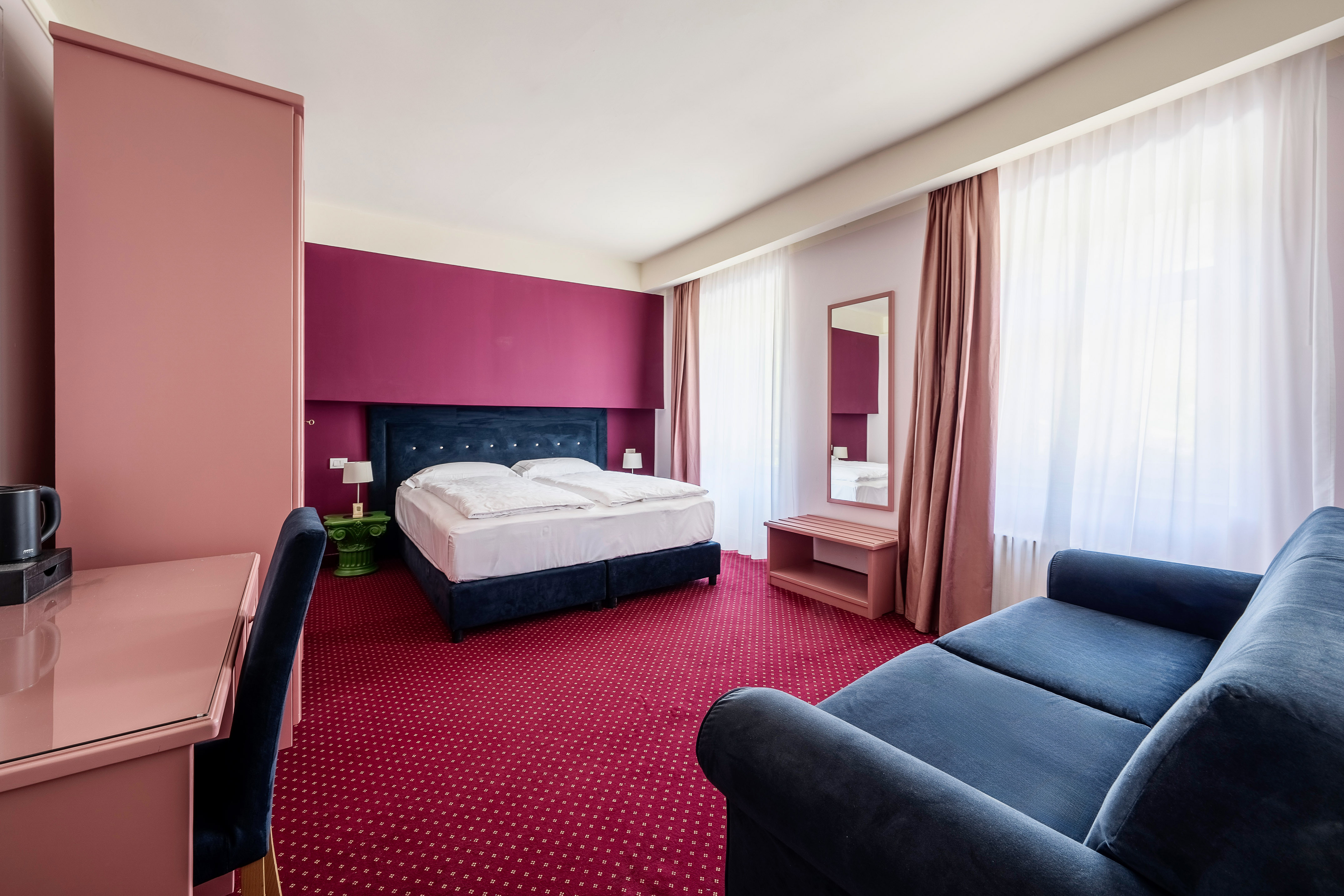 Vibrant hotel room: blue bed, red carpet, pink/magenta accent wall, blue sofa.