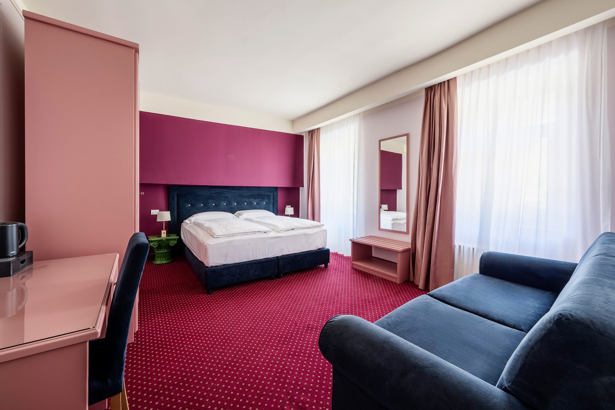 Vibrant hotel room: blue bed, red carpet, pink/magenta accent wall, blue sofa.