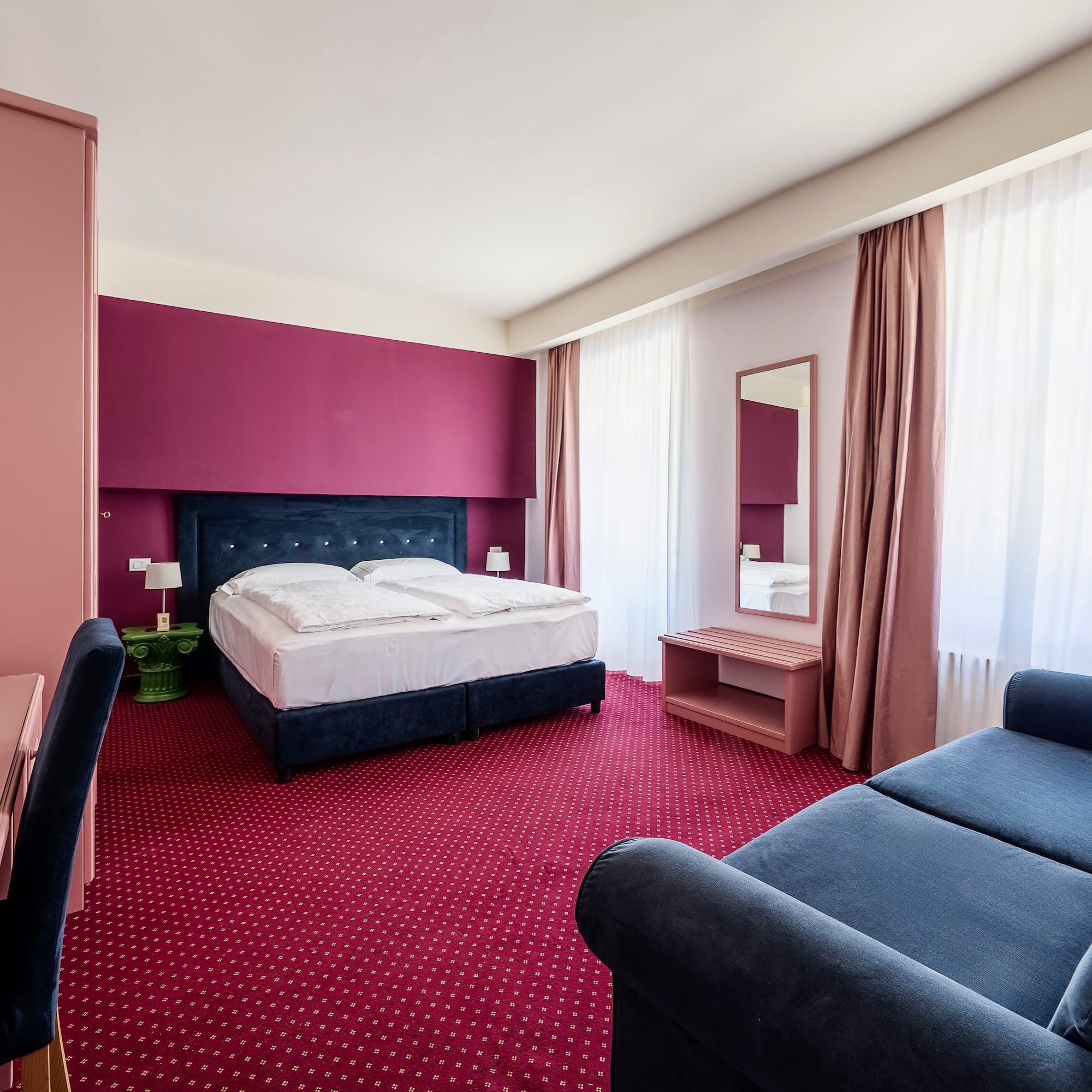 Vibrant hotel room: blue bed, red carpet, pink/magenta accent wall, blue sofa.