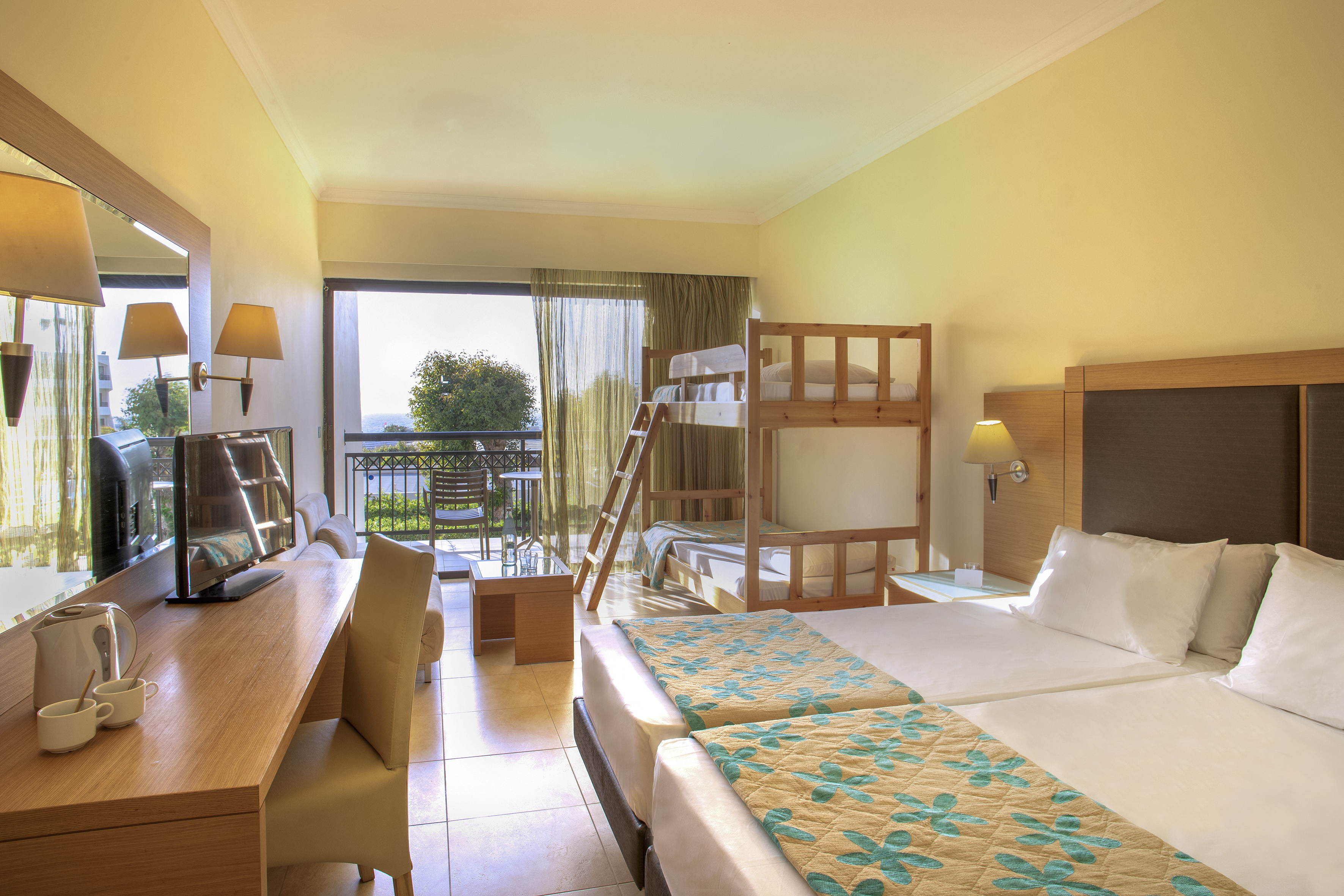 Bright hotel room with twin beds, bunk beds, desk, and balcony.