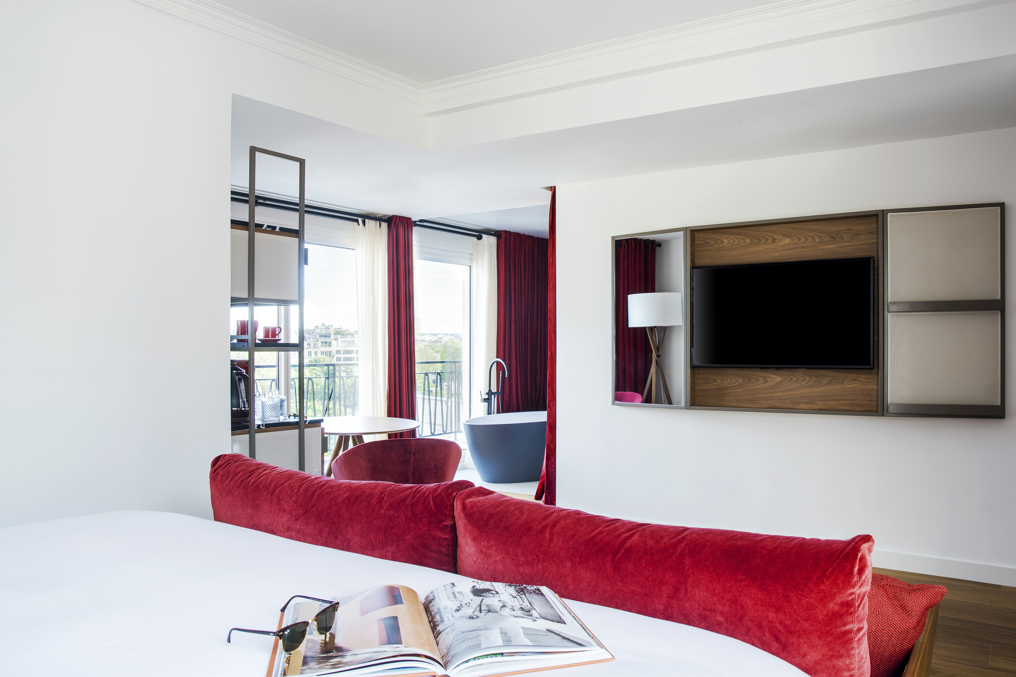 Modern hotel room with red bed, wall TV, balcony view, and freestanding tub.