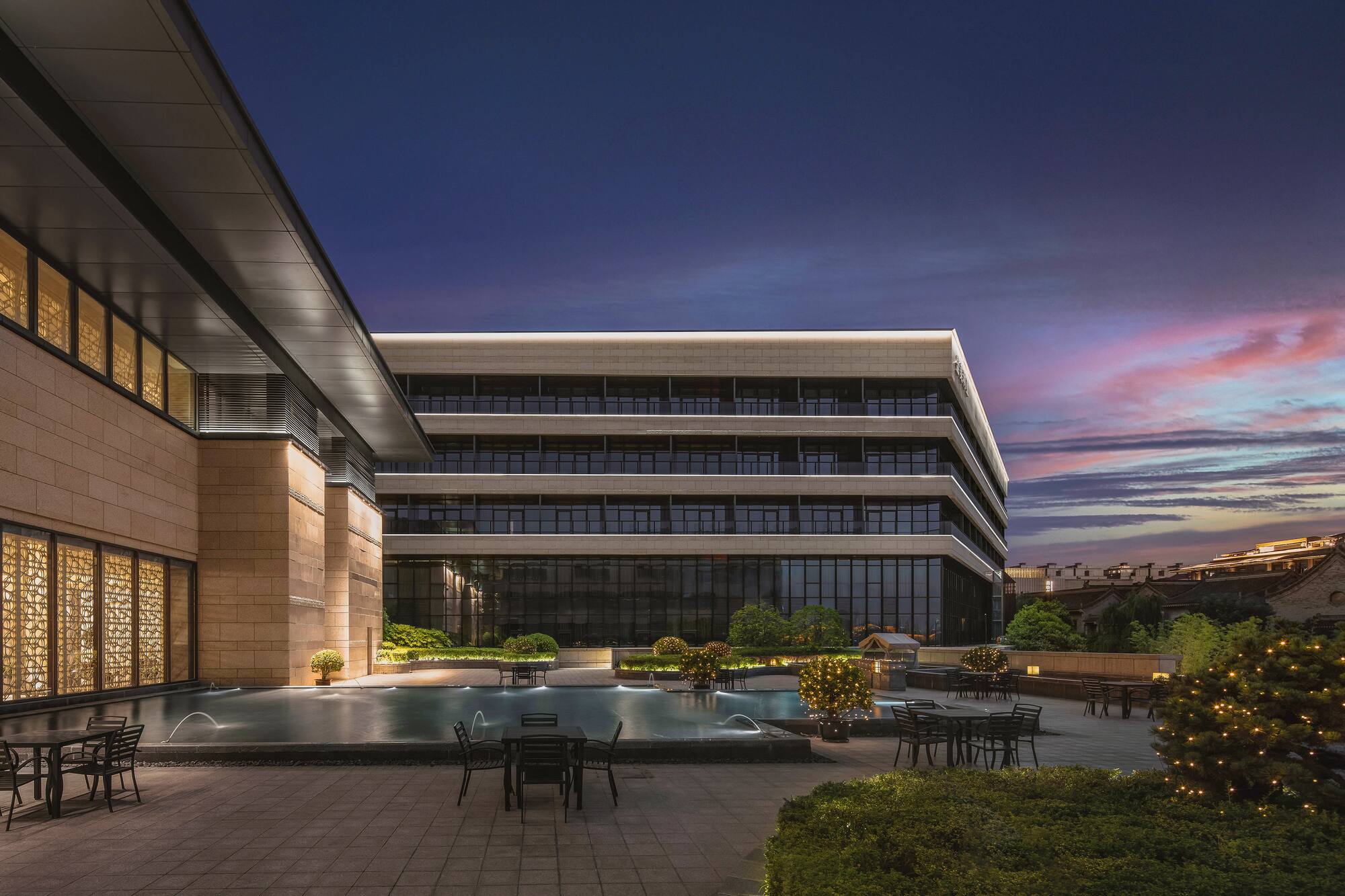 Modern building, illuminated patio, water feature, outdoor seating at sunset.