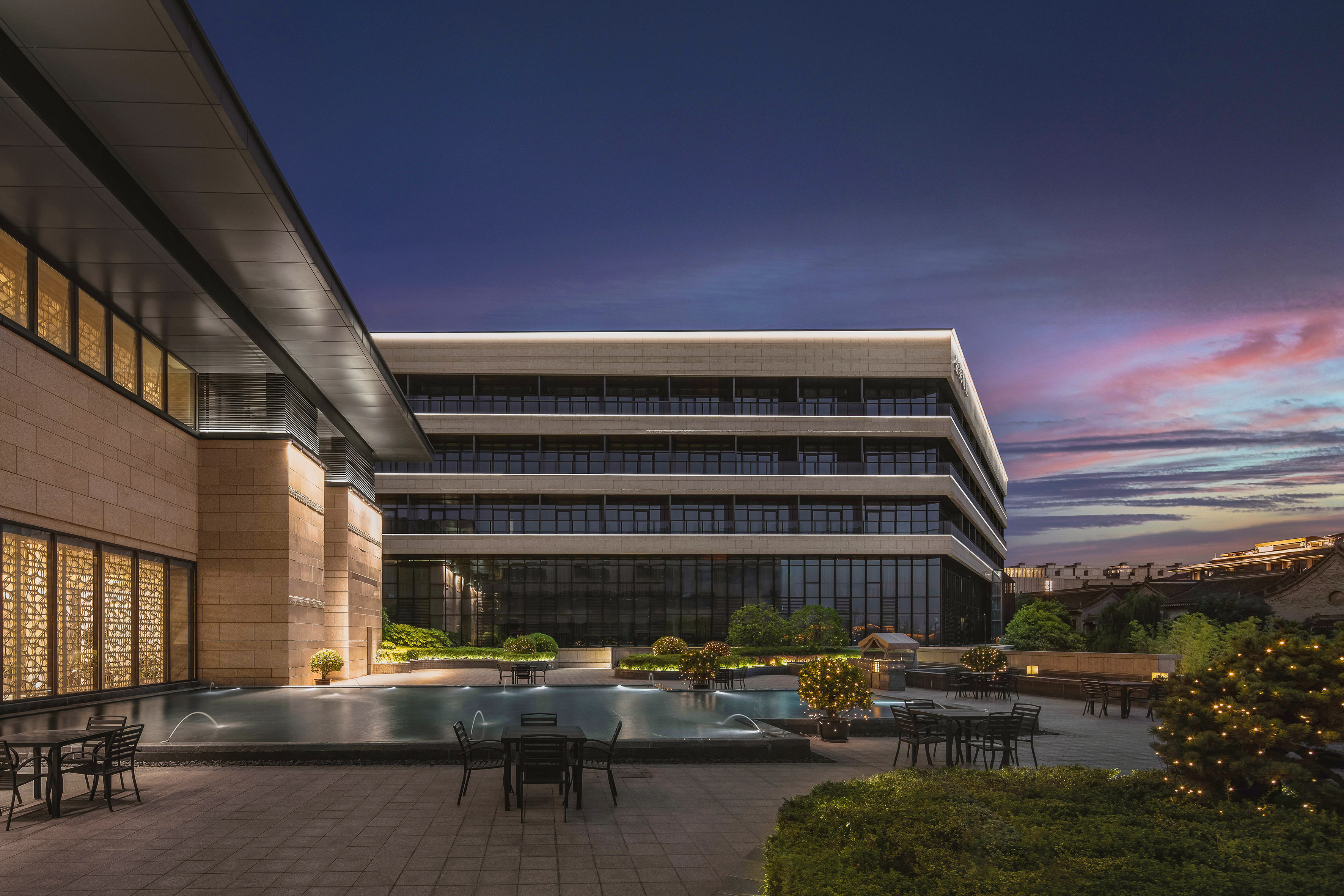 Modern building, illuminated patio, water feature, outdoor seating at sunset.