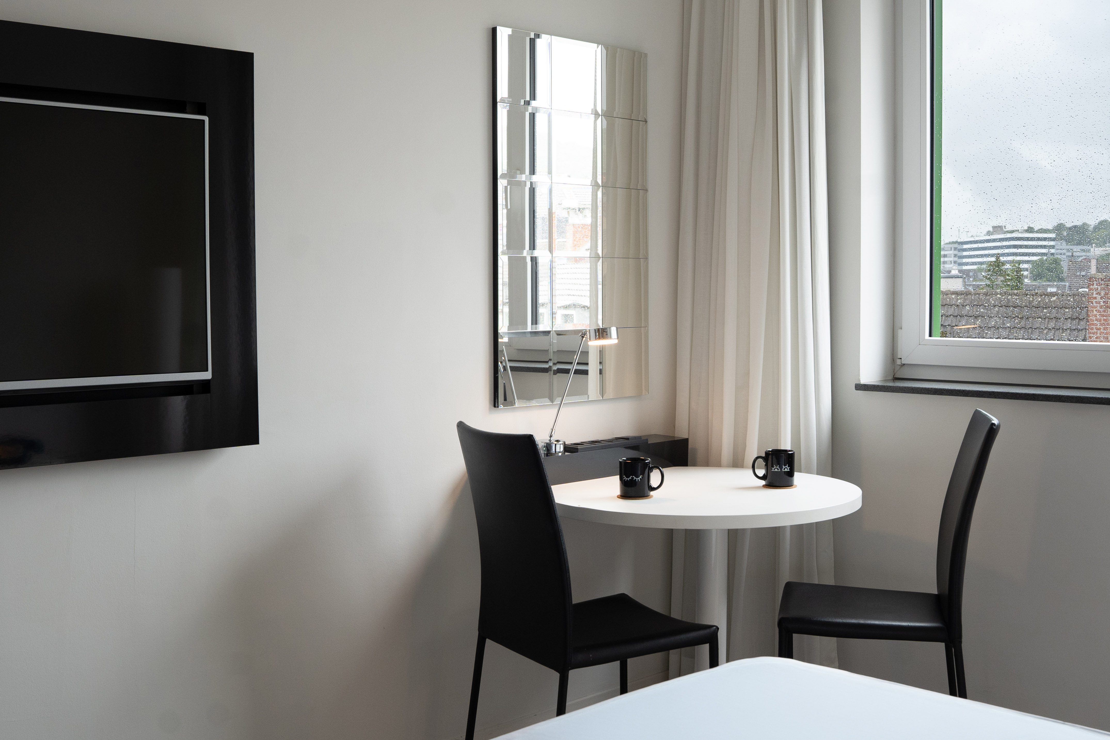 Modern hotel room with TV, round table, two chairs, mirror, and window view.