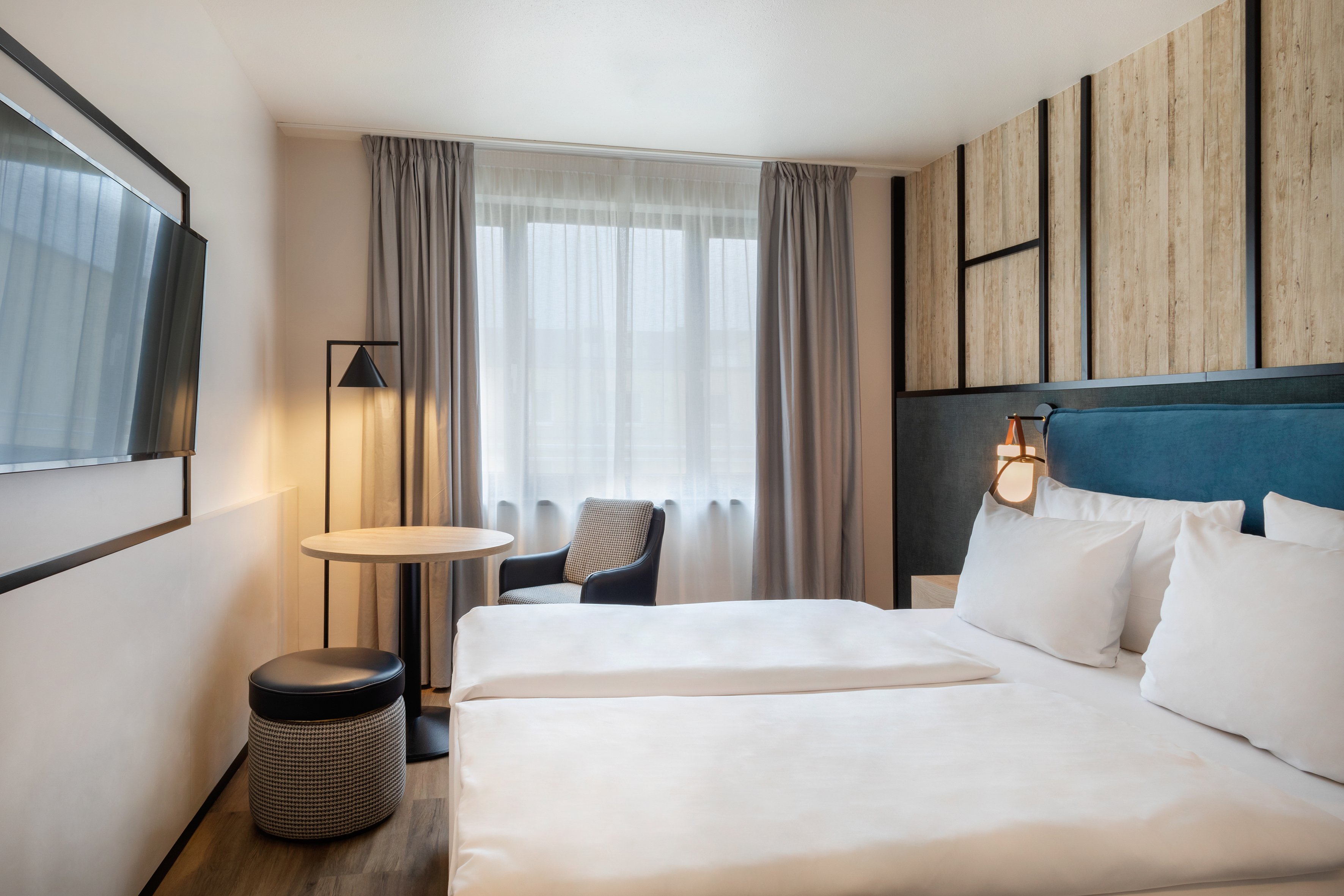 Modern hotel room with white bed, blue headboard, TV, and warm wood accents.