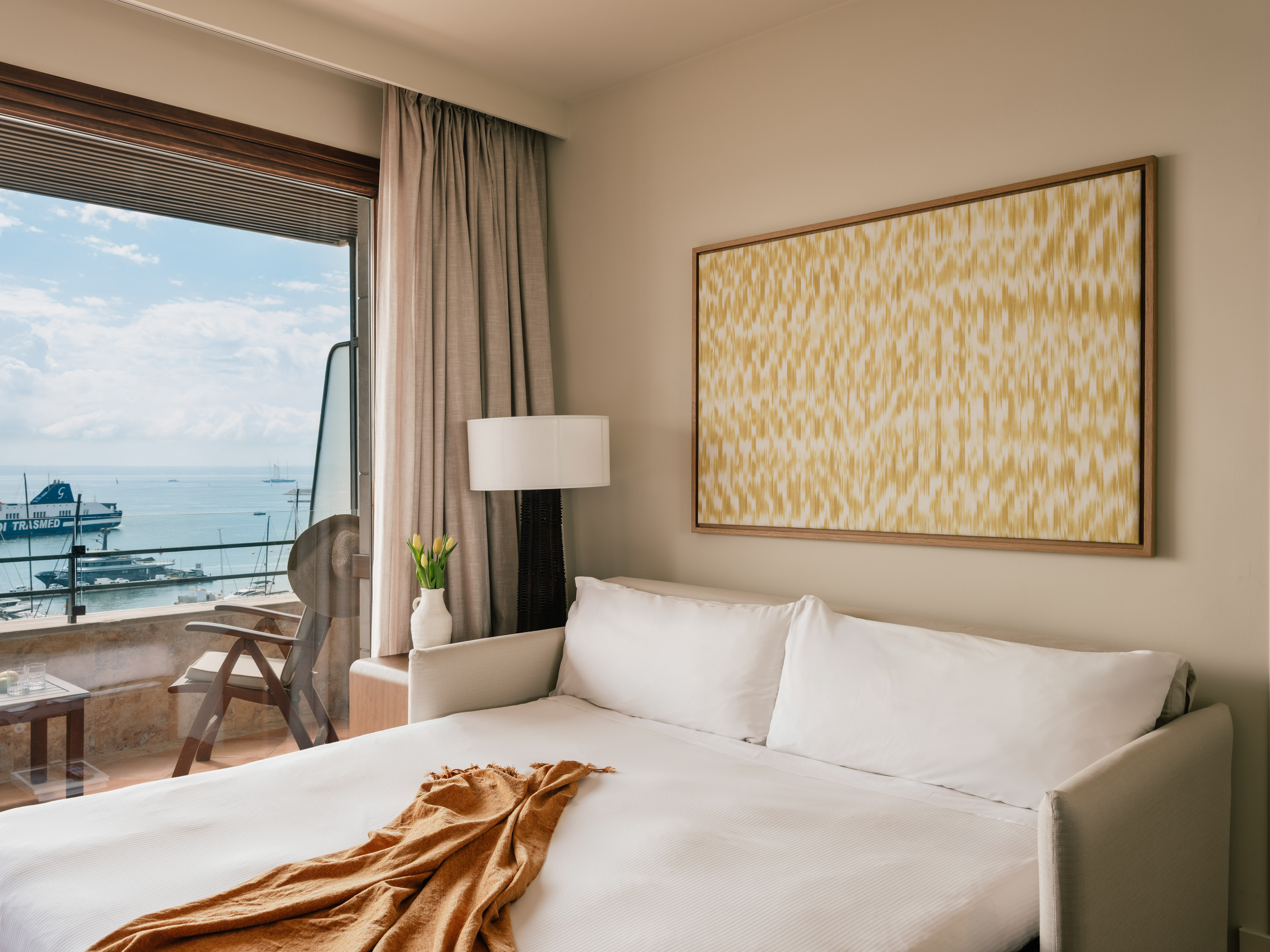 Hotel room with harbor view balcony, white sofa bed, and gold abstract art.