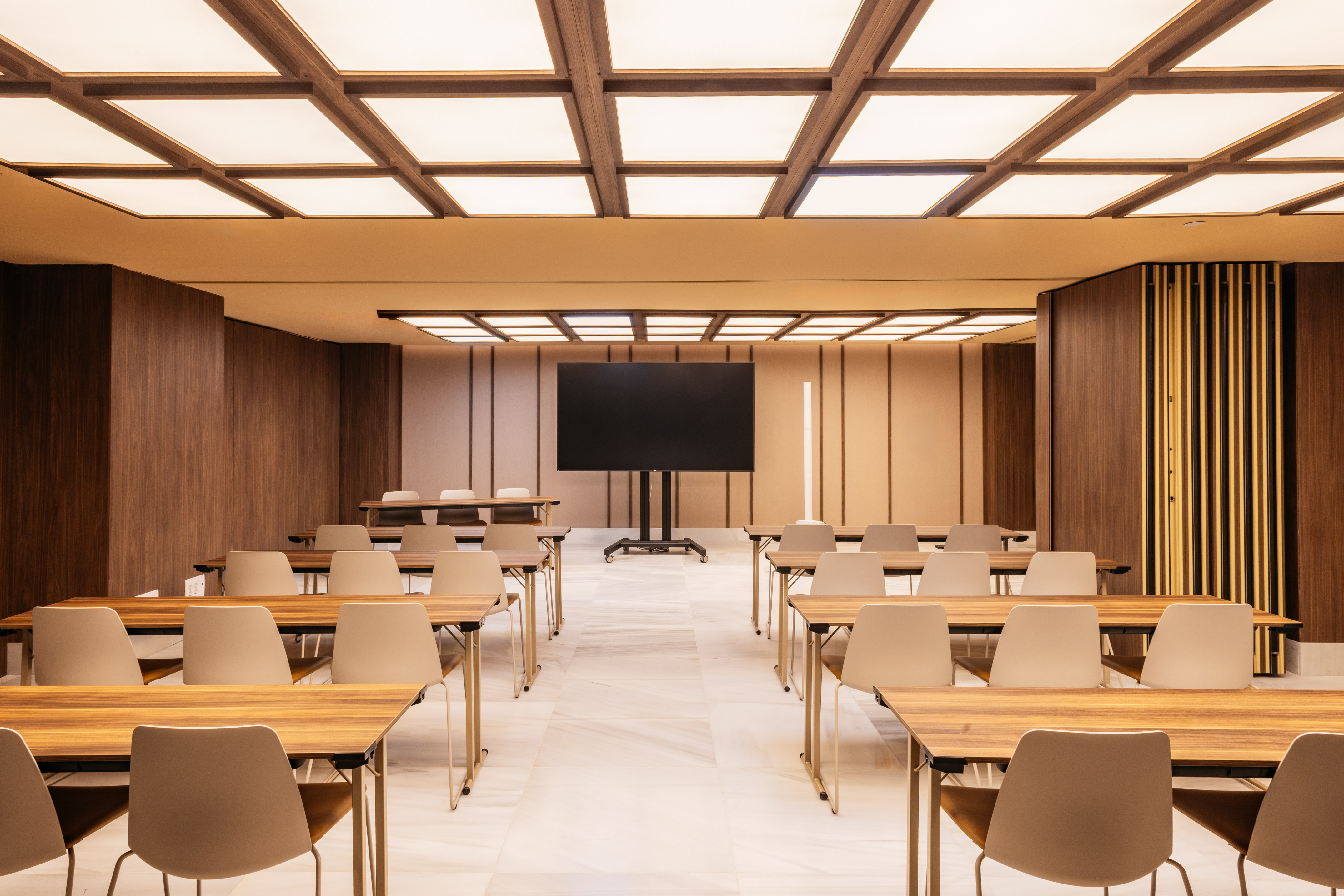 Modern conference room with tables, chairs, large screen, bright ceiling.