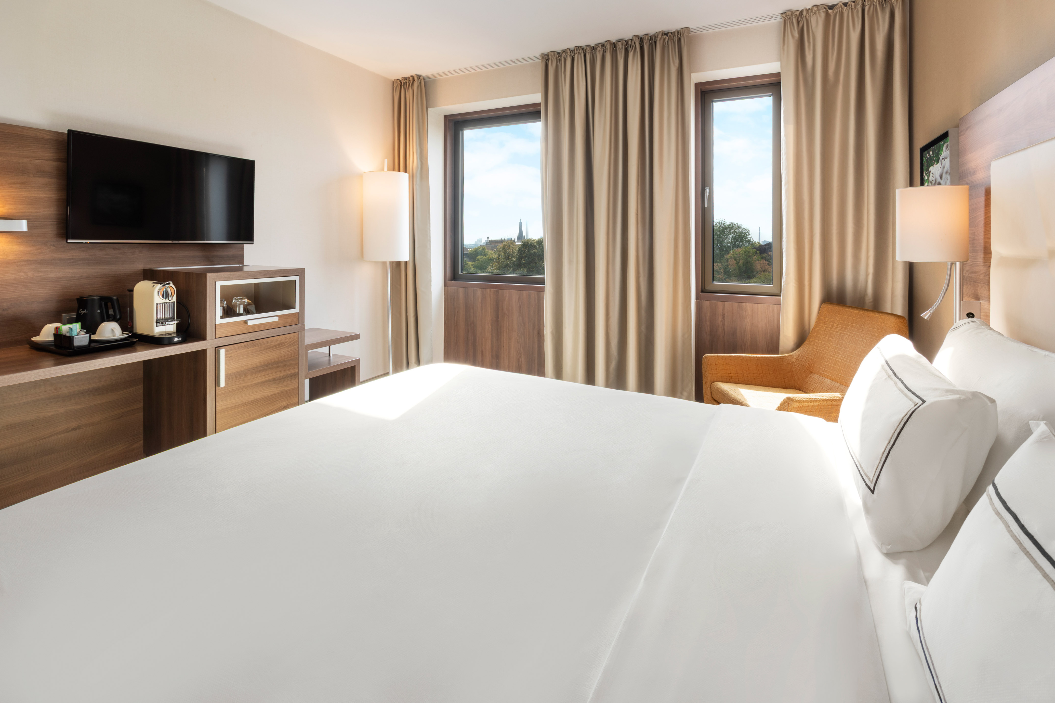 Modern hotel guest room with white bed, TV, coffee maker, and city view windows.
